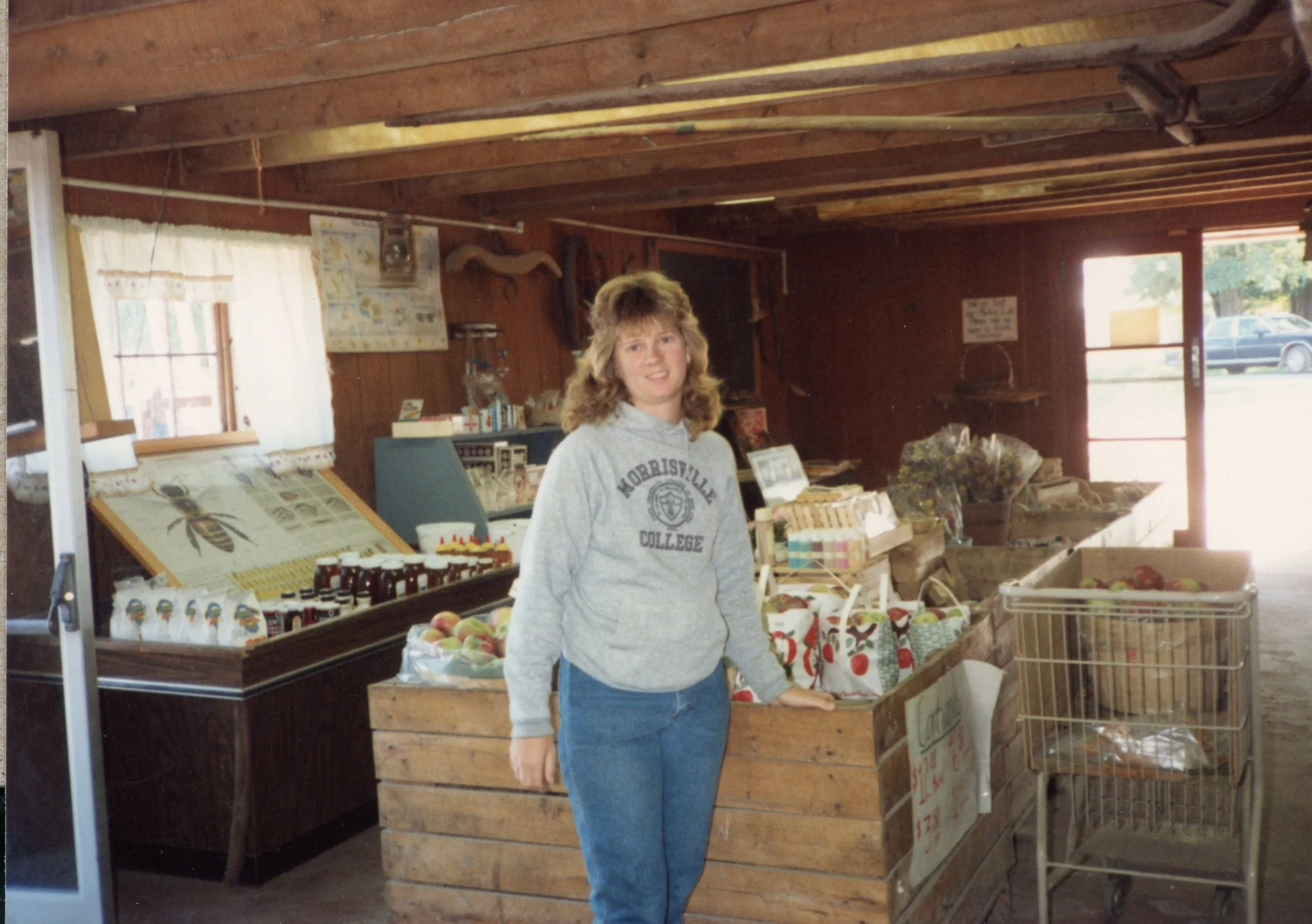 A young woman standing inside a rustic farm stand or market, surrounded by wooden crates of produce, with a shopping cart nearby. She is wearing a gray hoodie with 'Noblesville College' printed on it and blue jeans. The interior has wooden walls and ceiling, with a door leading outside, and various items displayed for sale.