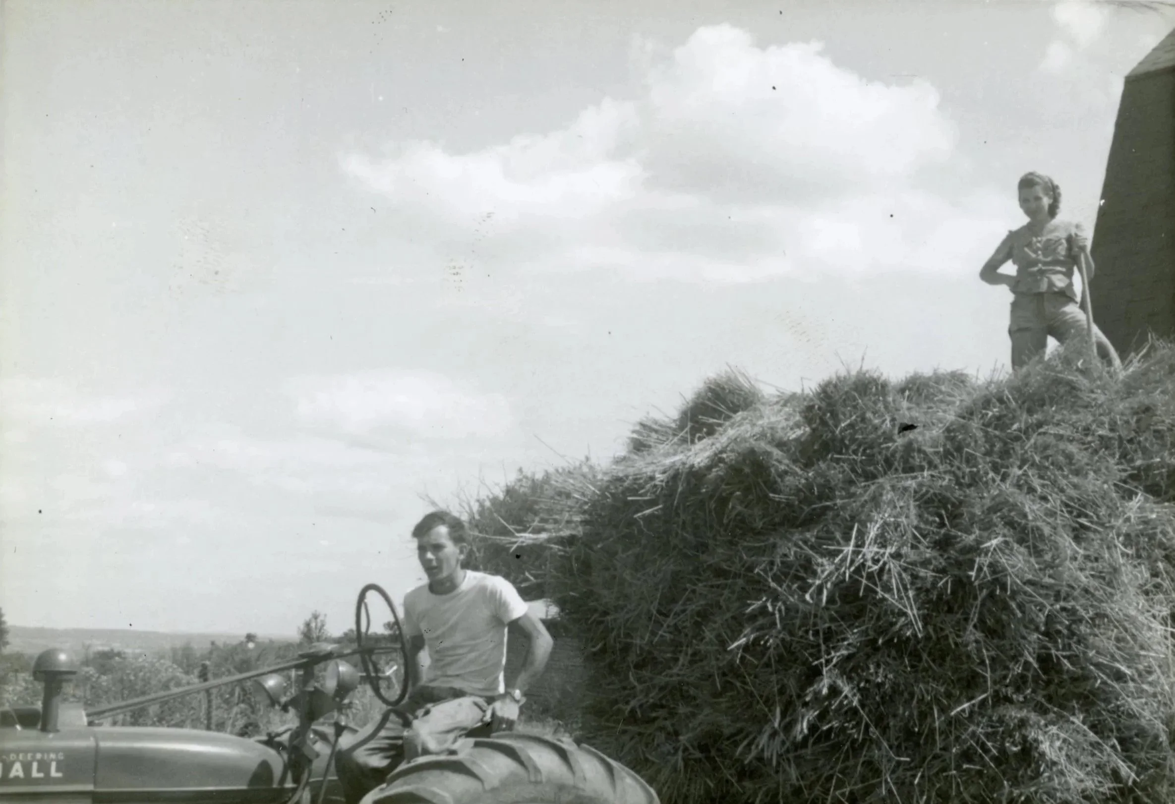 A man operating a tractor with a large load of hay, and a girl standing on top of the haystack on a farm.