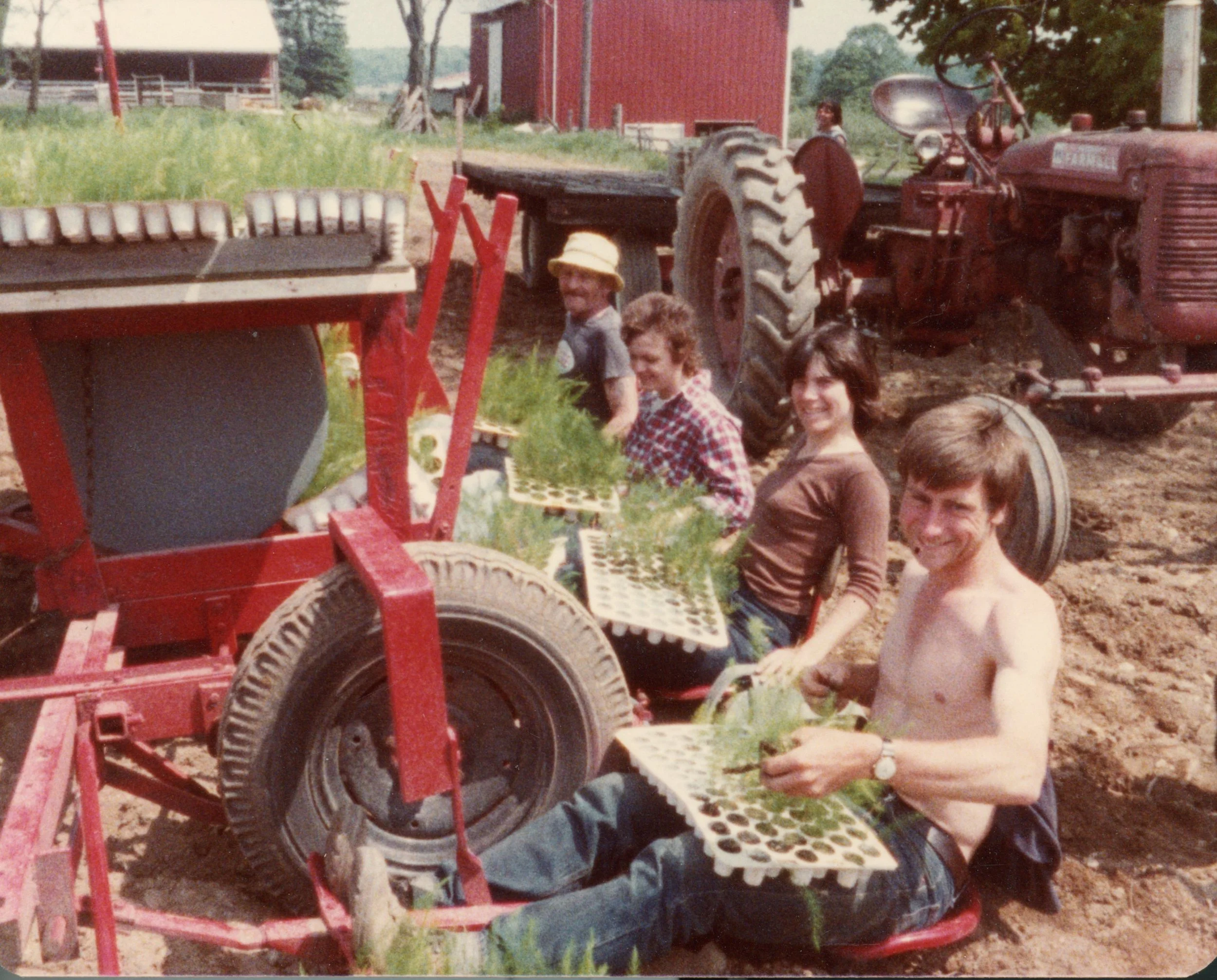 A group of people gardening outdoors, planting seedlings with trays in front of an old tractor on a farm.