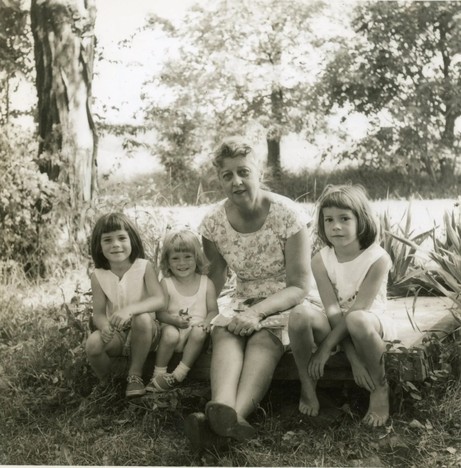 Four young girls and an older woman sitting outdoors on a wooden bench, surrounded by trees and plants, in black-and-white.