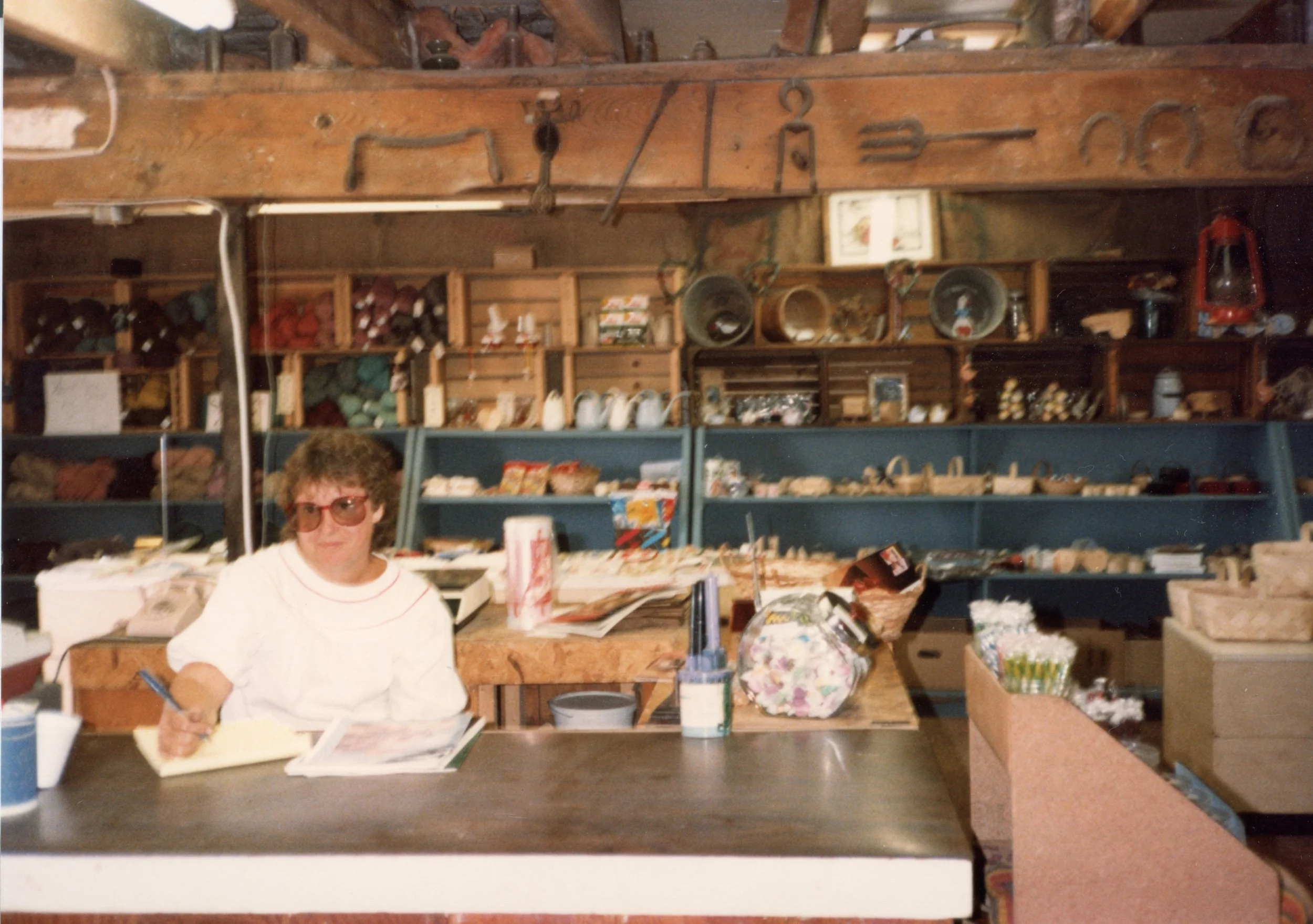 A woman sitting at a counter in a store, surrounded by shelves filled with various items such as yarn, jars, and knick-knacks, with a rustic wooden ceiling overhead.