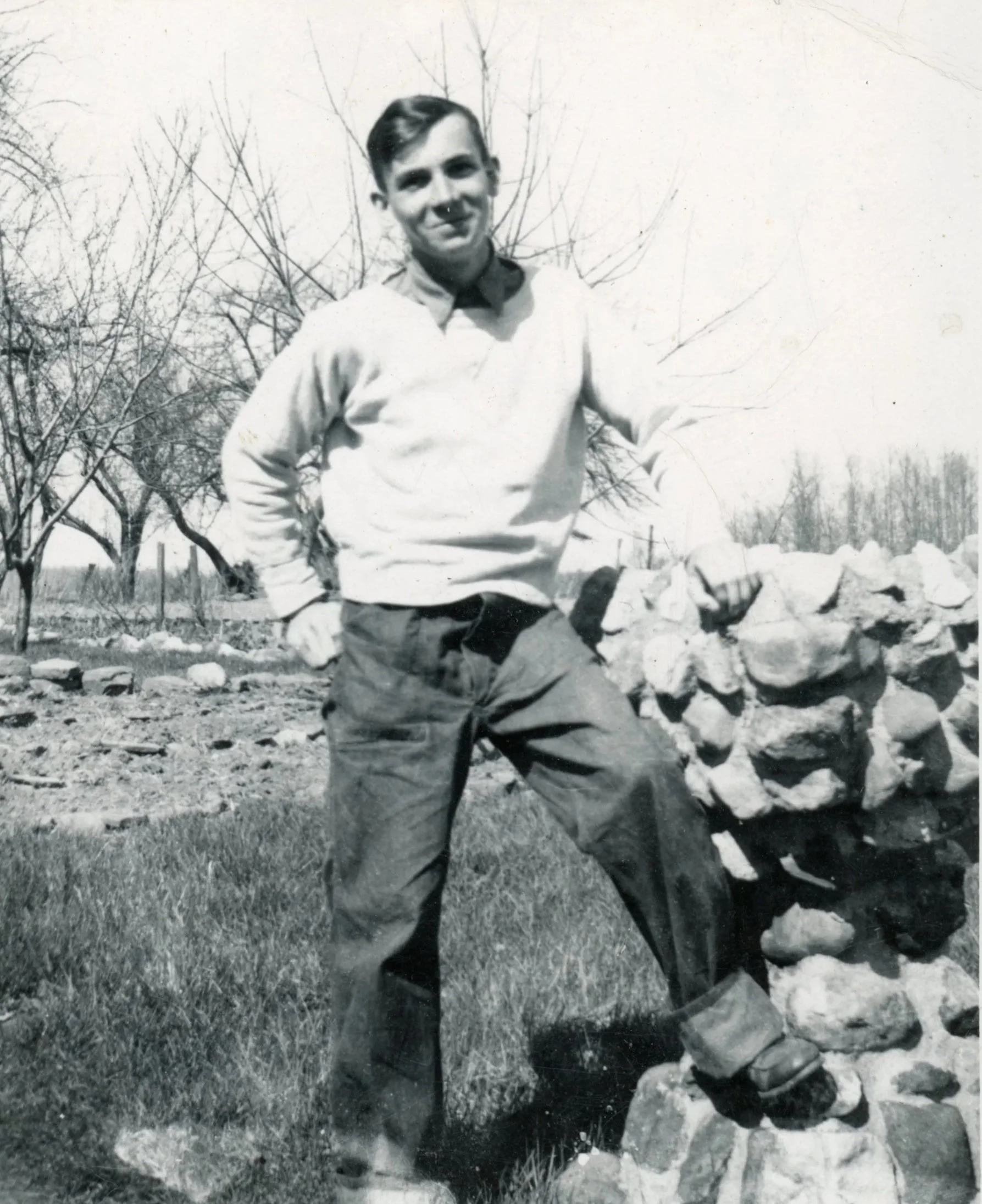 A young man with dark hair standing outdoors next to a low stone wall, wearing a light-colored sweater, dark pants, and boots, with leafless trees and a fence in the background.