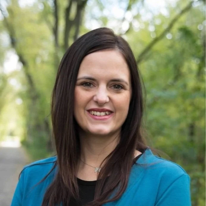 Woman long dark brown hair smiling while wearing black shirt with teal cardigan overtop. Green trees and forest blurred in the background.