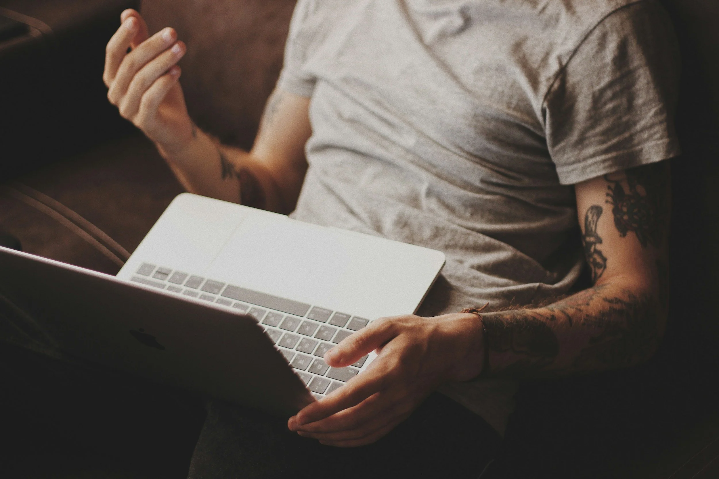 Man sitting down, wearing tan shirt and dark pants with arm tattoos.  Holding a gray laptop.
