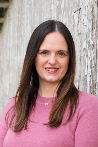 Smiling Woman with long dark brown hair in pink sweater in front of a weathered white chippy building.