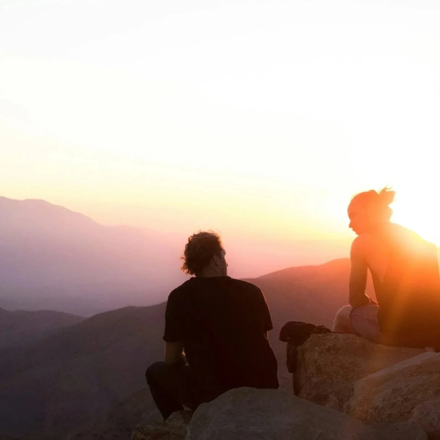 A man and woman sit on large rocks on the edge of a mountain looking at a purple, yellow and orange sunset.