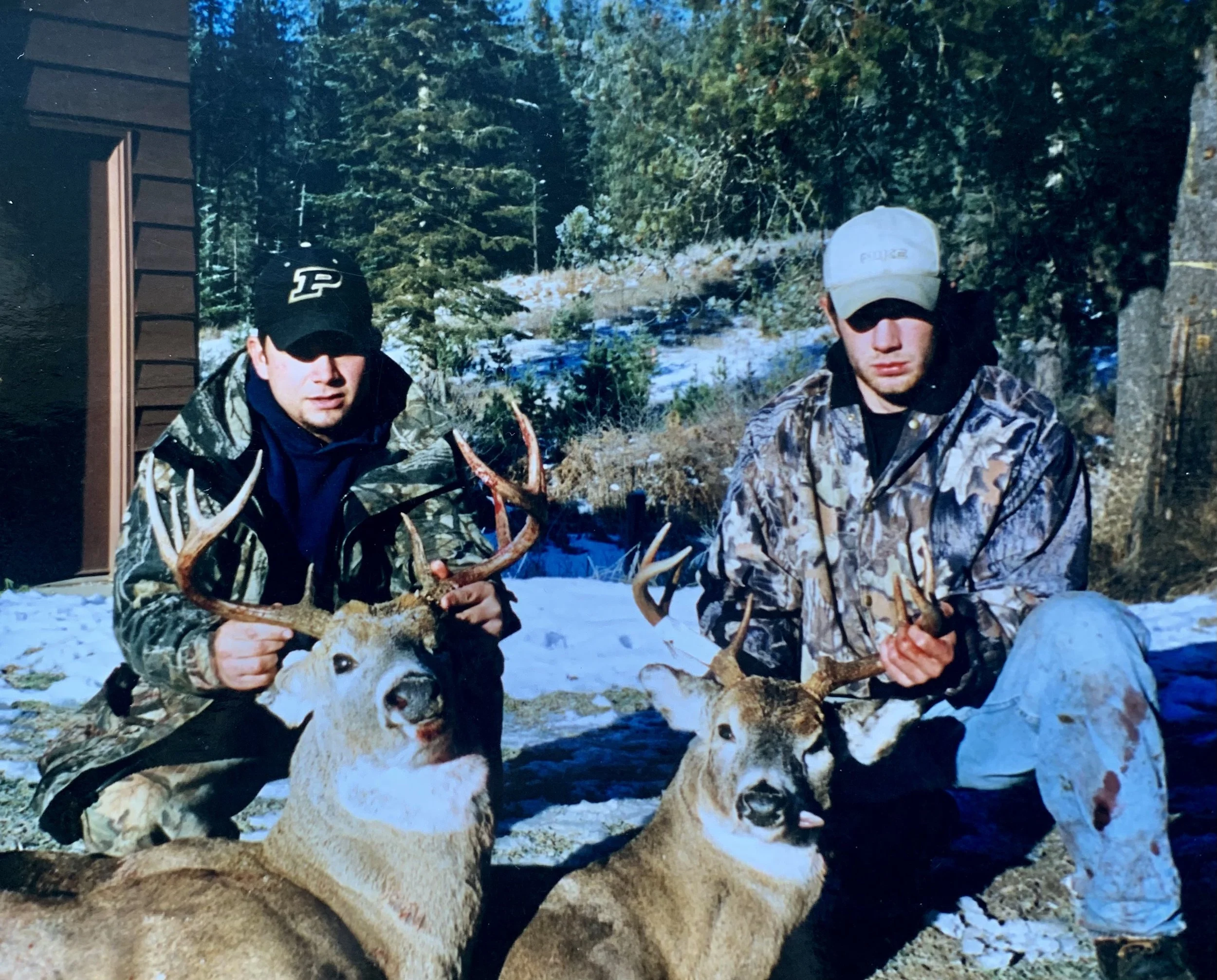 Two men outdoors in a snowy forest holding antlers from deer they have hunted, with one deer lying on the ground in front of each man.
