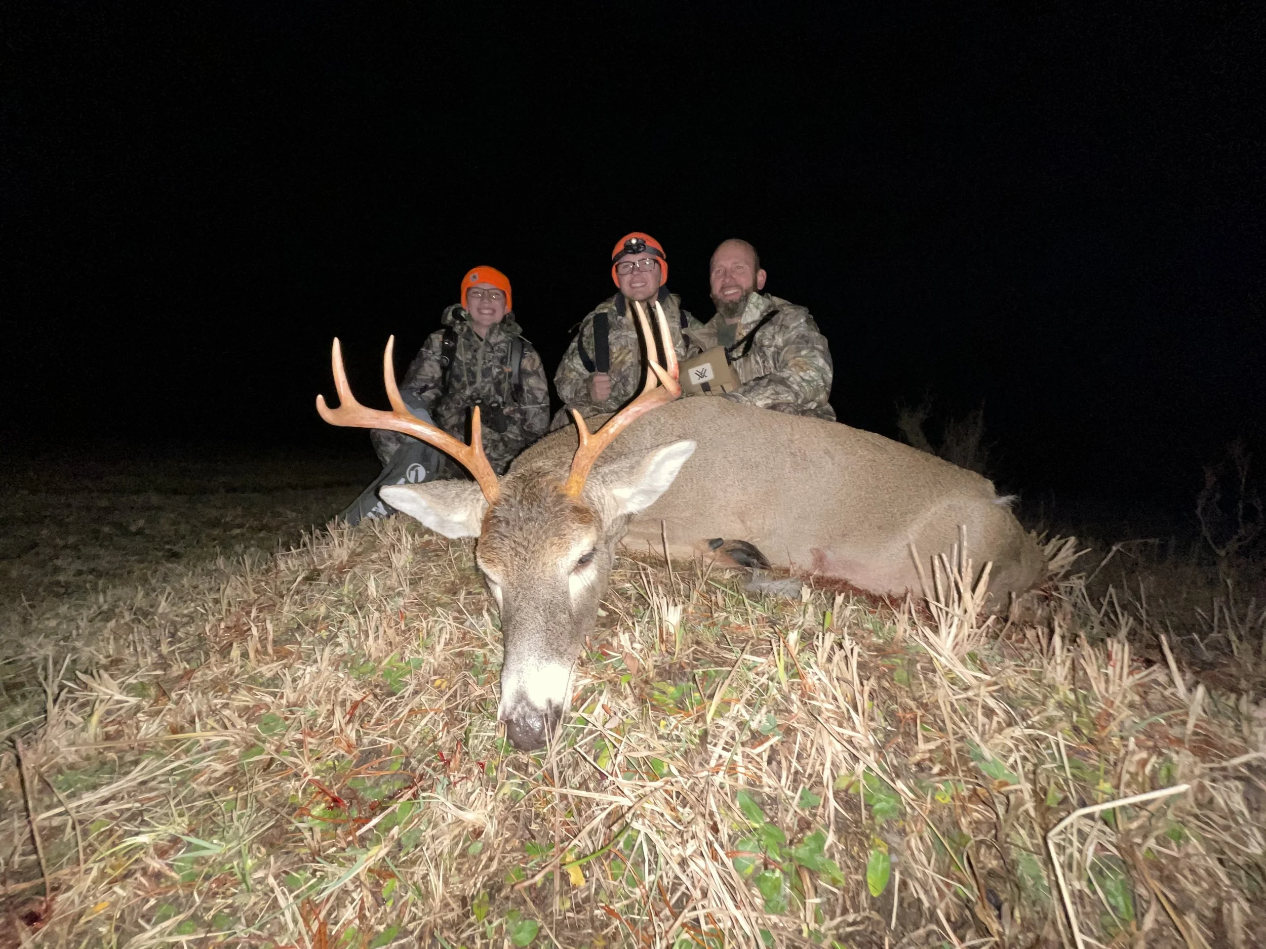 Three hunters in camouflage clothing and orange hats posing behind a large, freshly hunted deer with antlers lying on the ground at night.