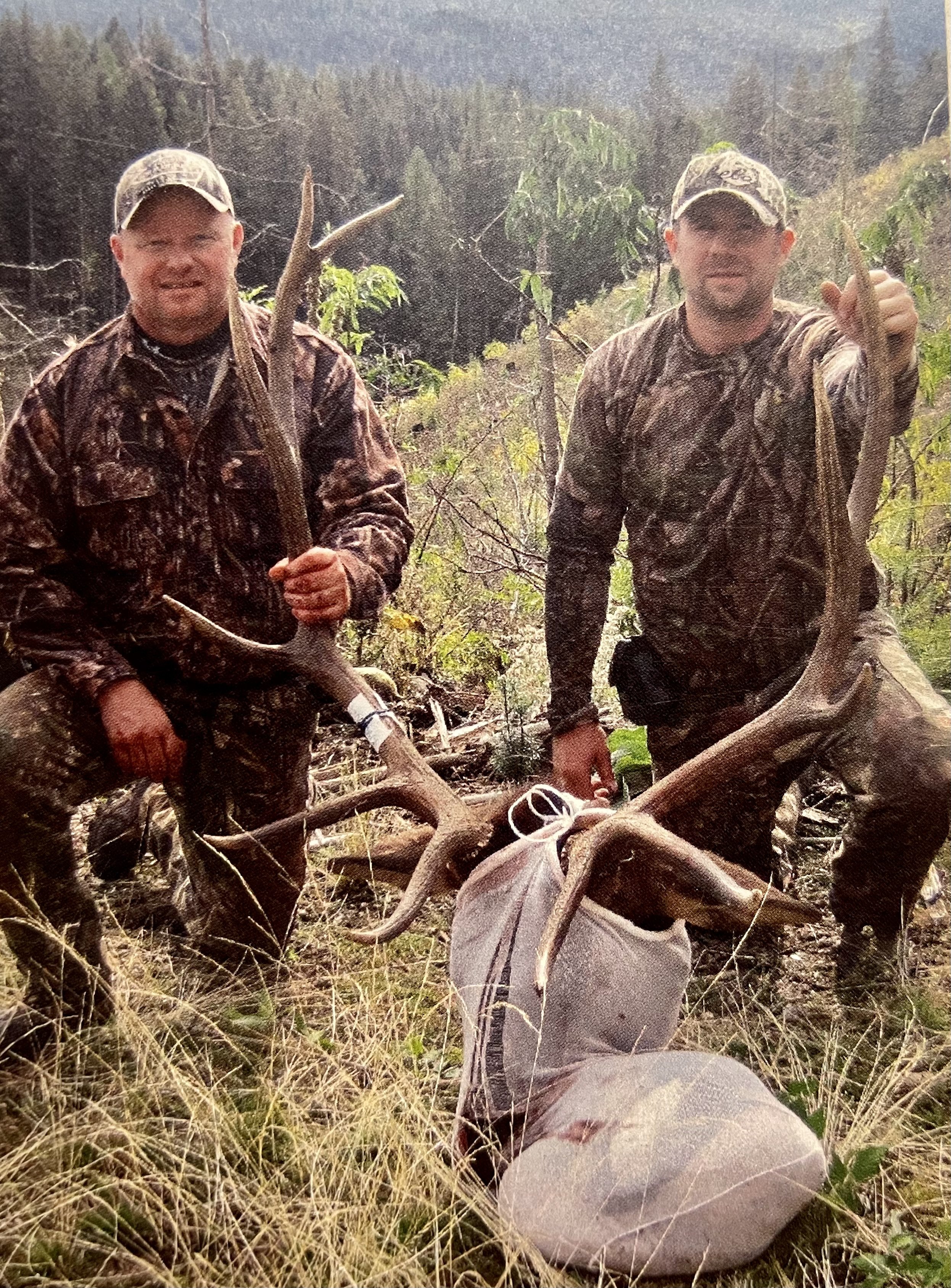 Two hunters in camouflage clothing holding large antlers, standing in a natural outdoor setting, with one of the antlers displayed in a bag on the ground.
