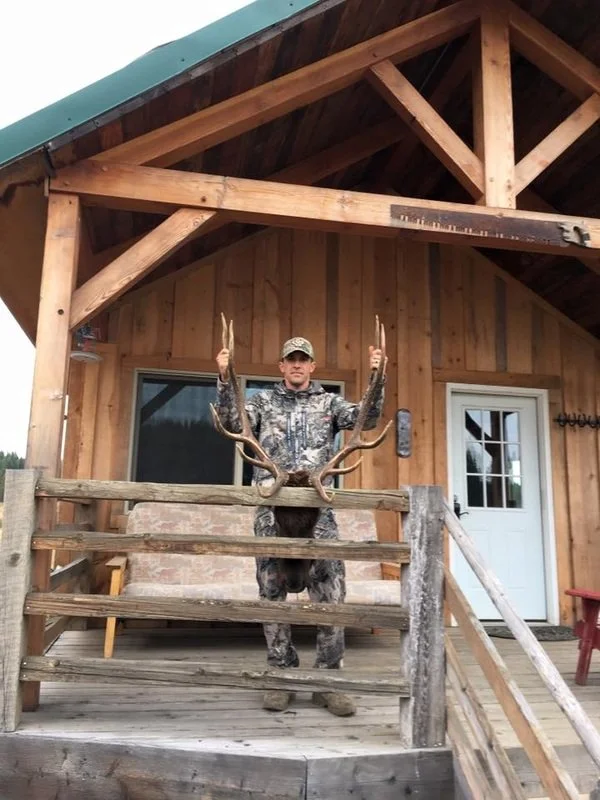 A man in camouflage clothing holding large antlers standing on the porch of a wooden cabin.