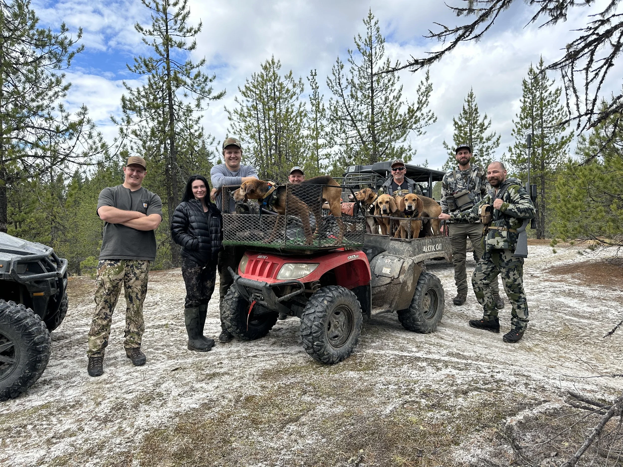 Group of people on a dirt trail in forest, standing around a red utility vehicle loaded with hunting dogs, with trees and cloudy sky in background.