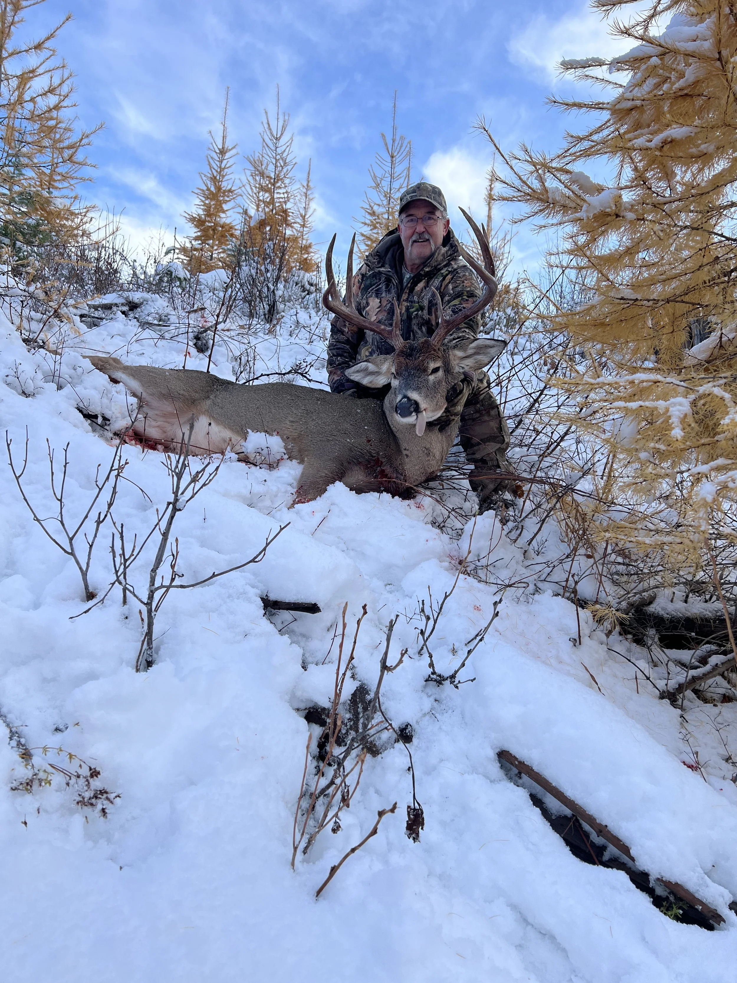 A man dressed in camouflage hunting gear kneels in snow-covered forest, posing with a large deer he has hunted, which has antlers and a visible tongue.