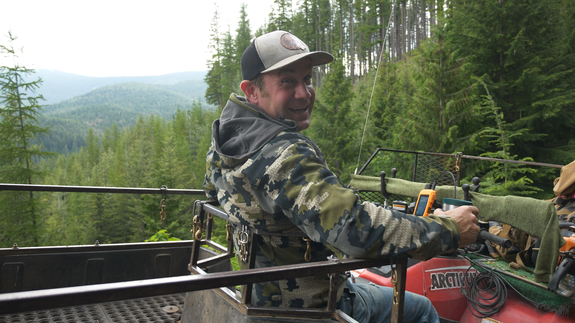 A man smiling while riding an off-road vehicle through a forested mountainous area with tall green trees and distant mountains in the background.