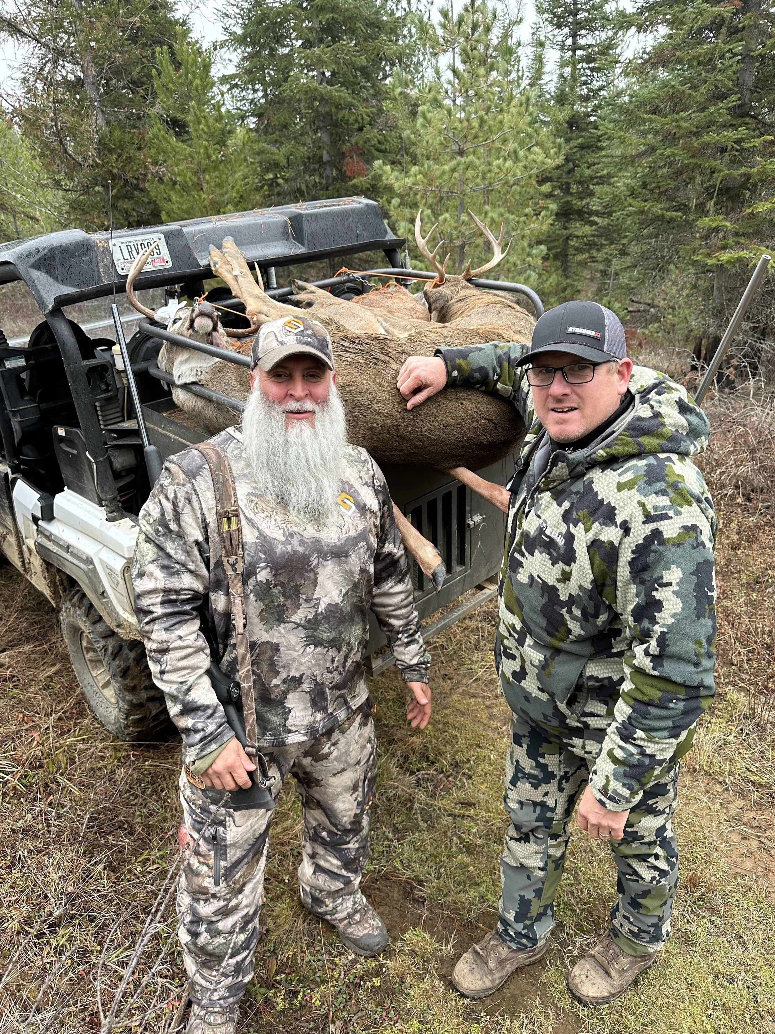 Two men in camouflage clothing standing beside a golf cart with a large elk and a smaller animal, likely a mountain goat or similar, on it in a wooded area.
