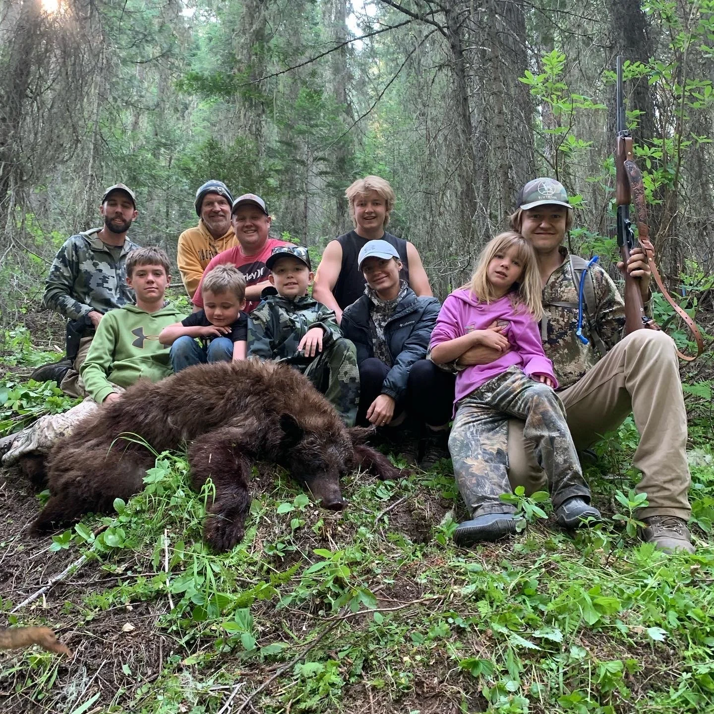 A group of people, including children and adults, posing in a forest with a dead bear in front of them. One man is holding a rifle.