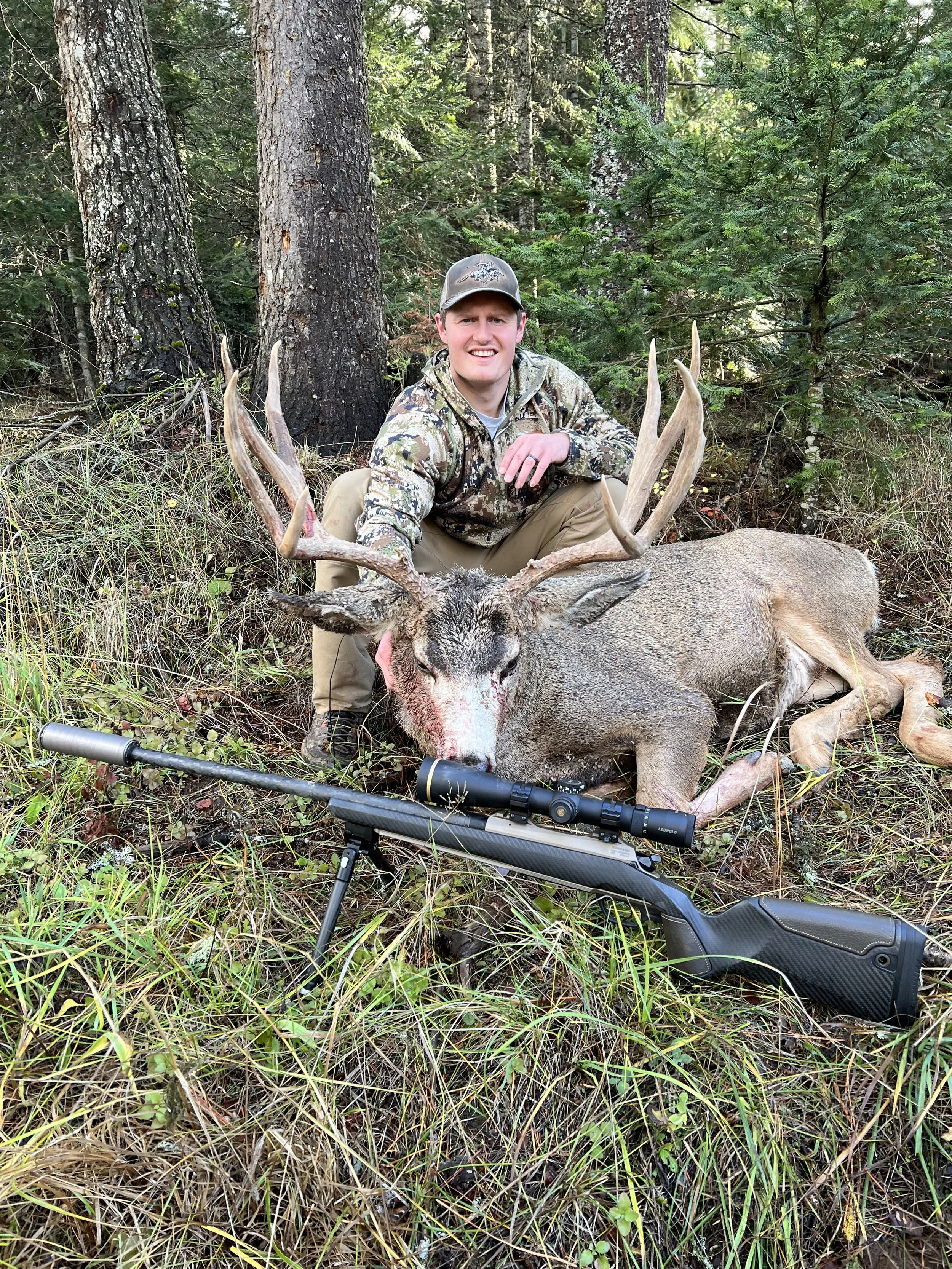 A man in camouflage clothing is kneeling behind a large deer with antlers, in a forest setting, with a rifle and scope in front of him.