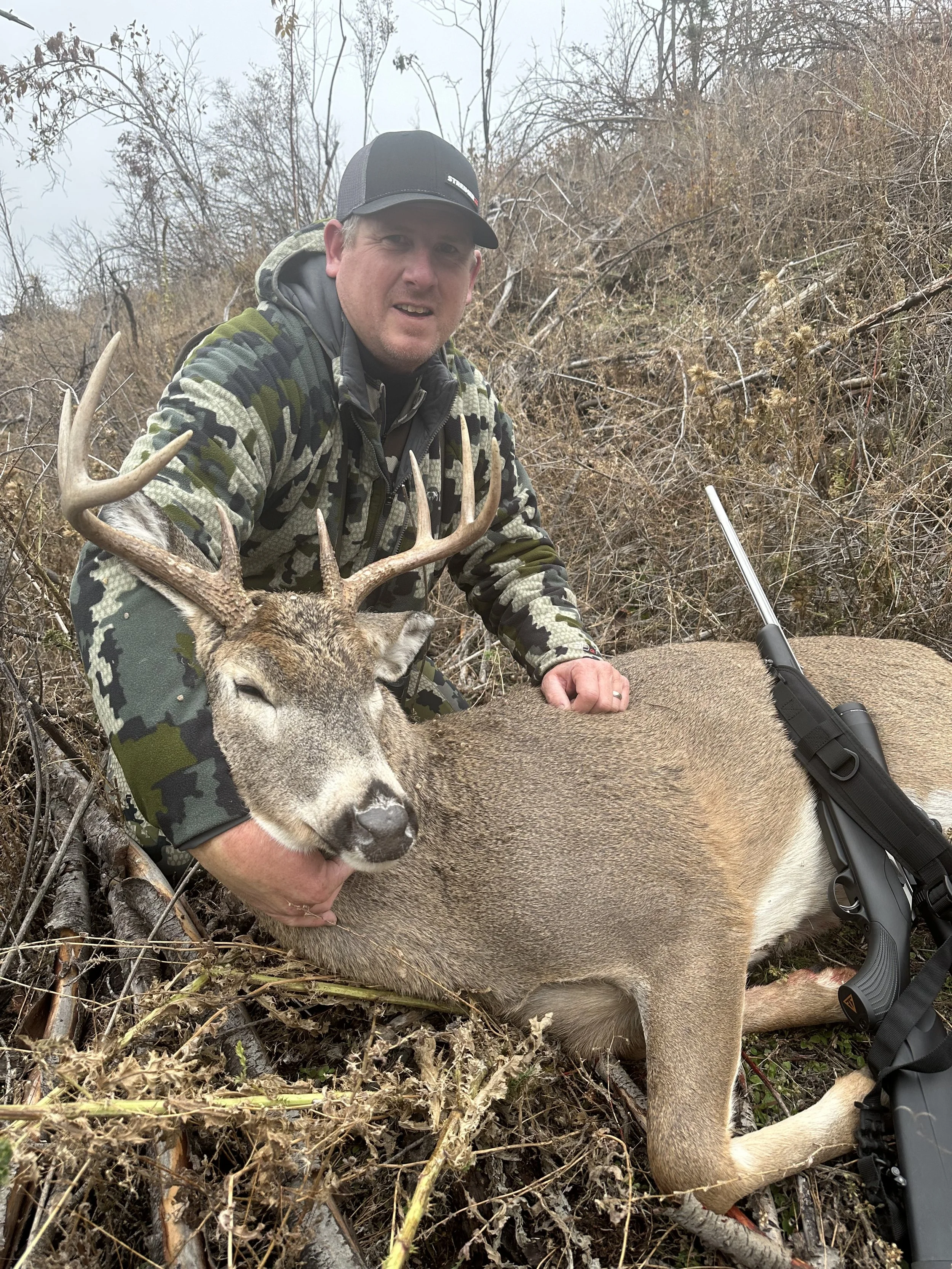 A man in camouflage clothing kneels on the ground holding a large deer with antlers. The man is wearing a black cap and appears to be in a wooded area with dry grass and branches.