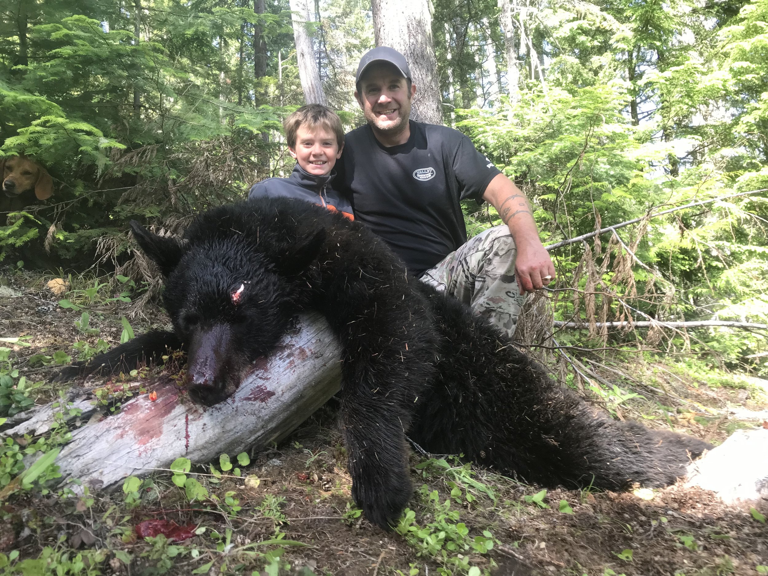Two smiling men and a boy kneel outdoors in a wooded area near a fallen tree with a large dead bear laying on it, with blood on its face and snout. One of the men holds a stick, and a dog peeks from the background among green trees and bushes.