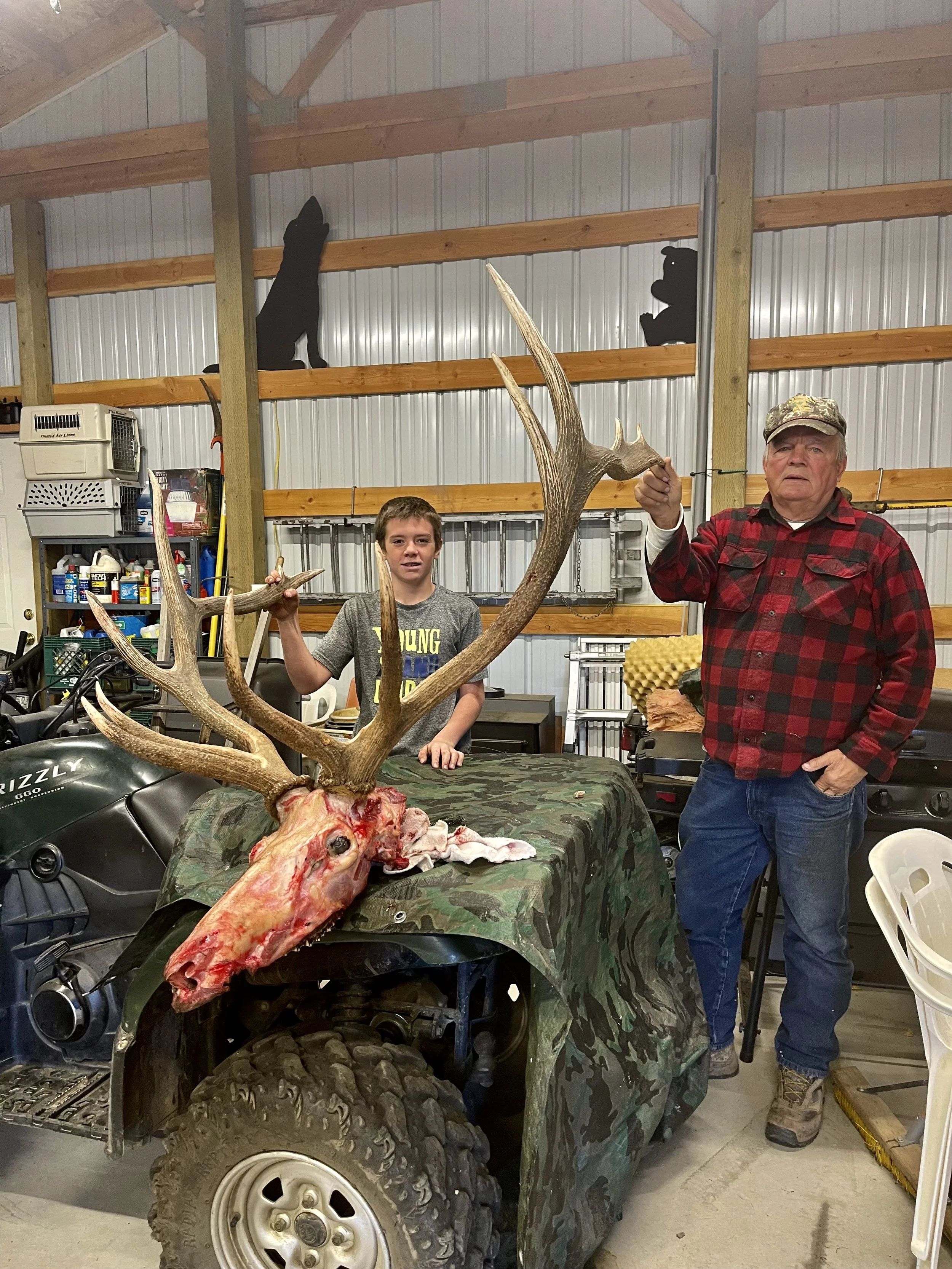A young boy and an older man standing in a garage next to a mounted deer head with large antlers. The young boy is holding the antlers, and the older man is holding a small object. The garage has shelves with various items, and there are silhouettes 