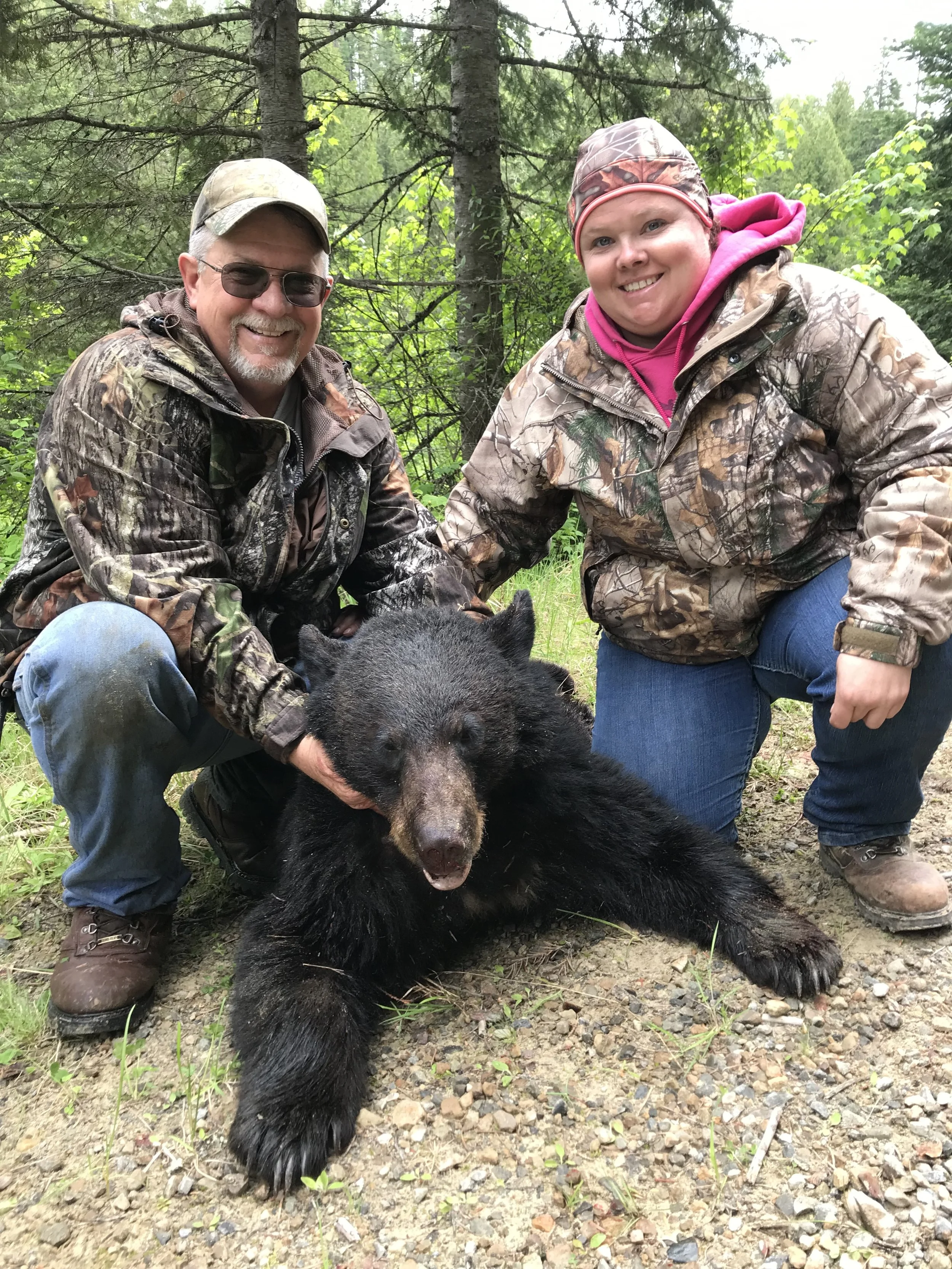 Two people, a man and a woman, in camouflage outdoor clothing posing with a large black bear cub in a forest setting.