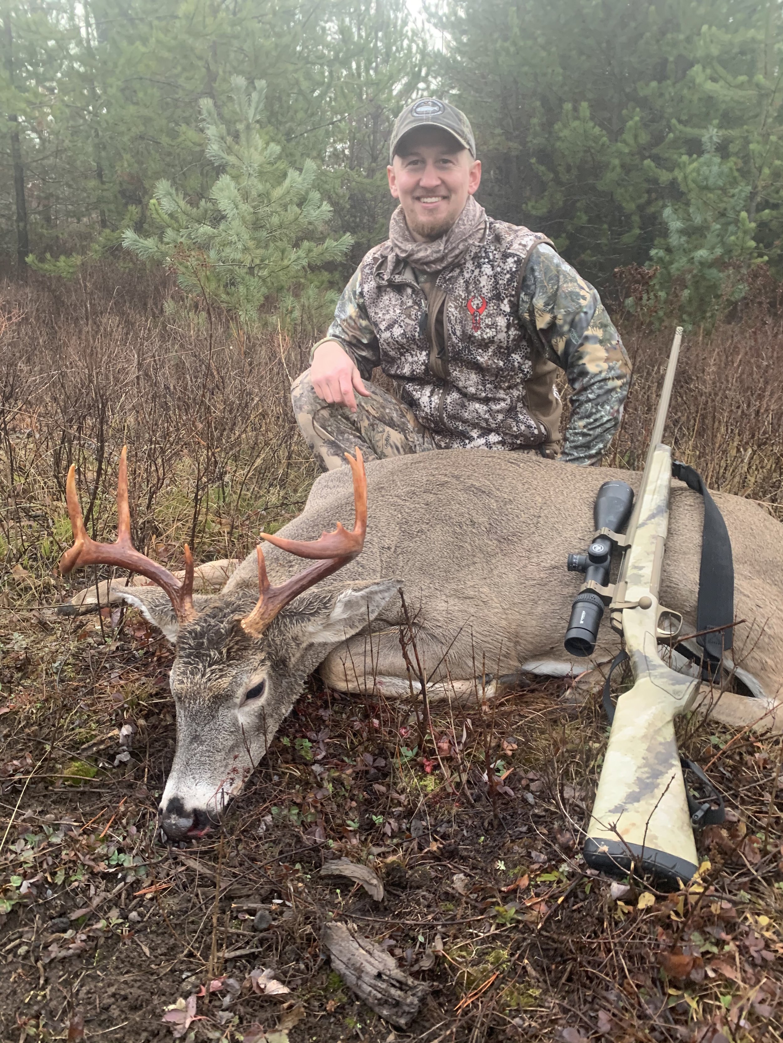 A man in camouflage hunting gear kneeling next to a large deceased deer with antlers, a scoped rifle resting on the deer, in a forested area.
