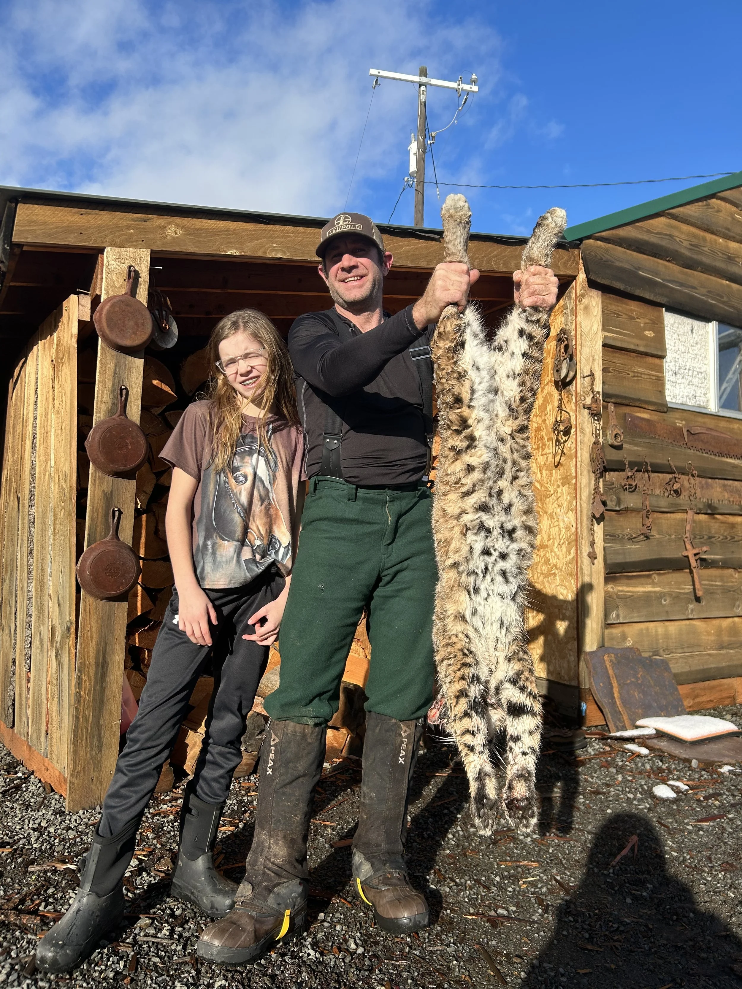 A man and a young girl standing outside a wooden shed, holding up a large mounted animal pelt, possibly a bobcat or lynx. The man is wearing green pants, boots, and a cap, while the girl is dressed in black pants and boots, smiling at the camera. The