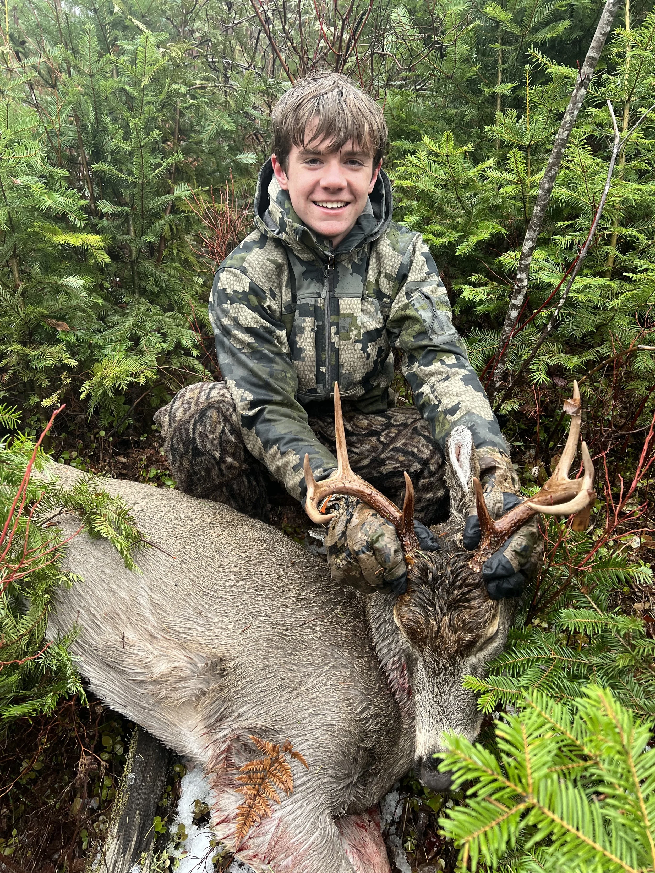 A young man dressed in camouflage hunting gear kneels among green foliage, holding the antlers of a large, deceased elk with gray fur and blood on its belly, in a forest setting.