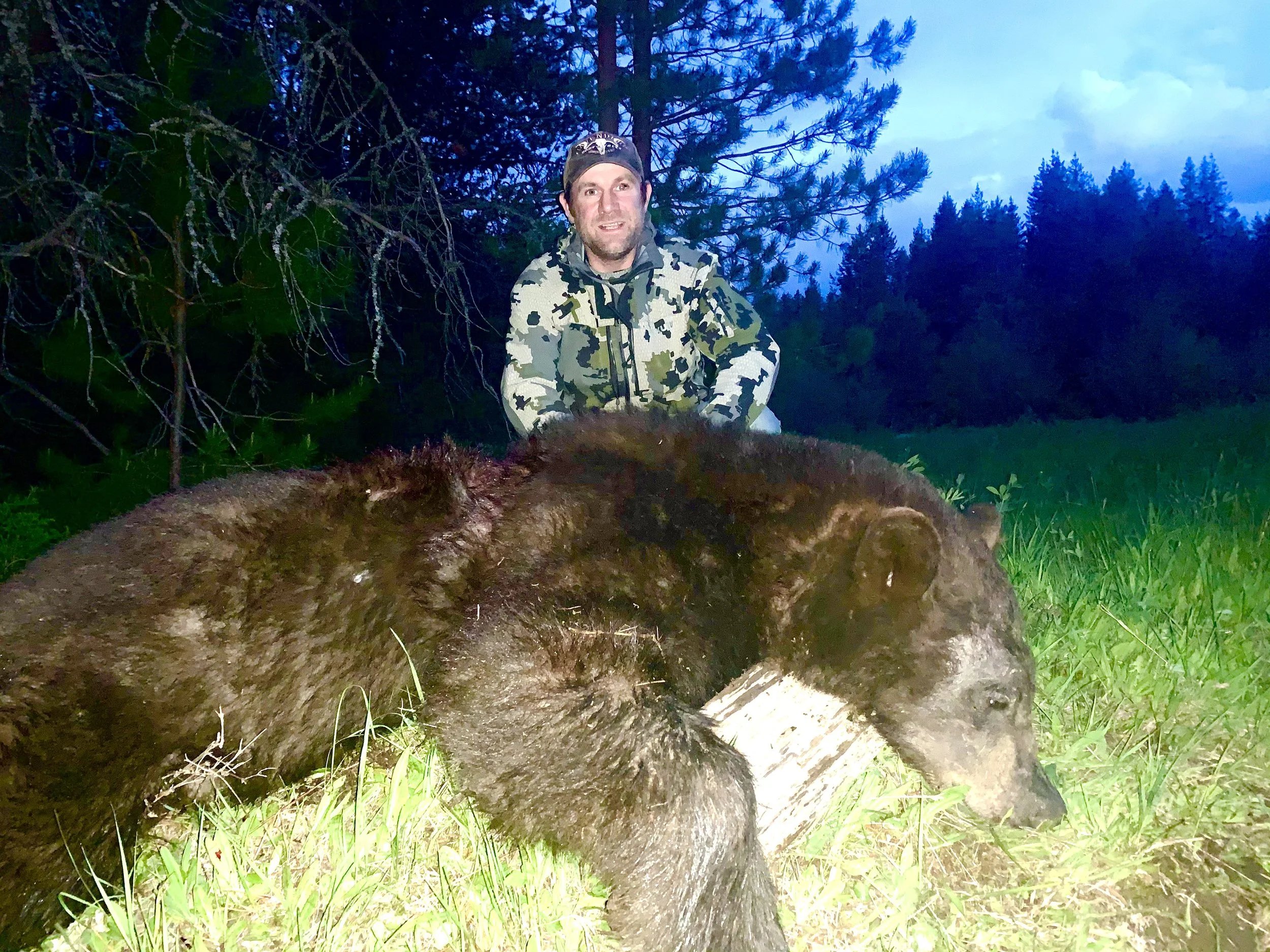 A man in camouflage clothing posing behind a large, dead bear in a forested area during dusk or dawn.