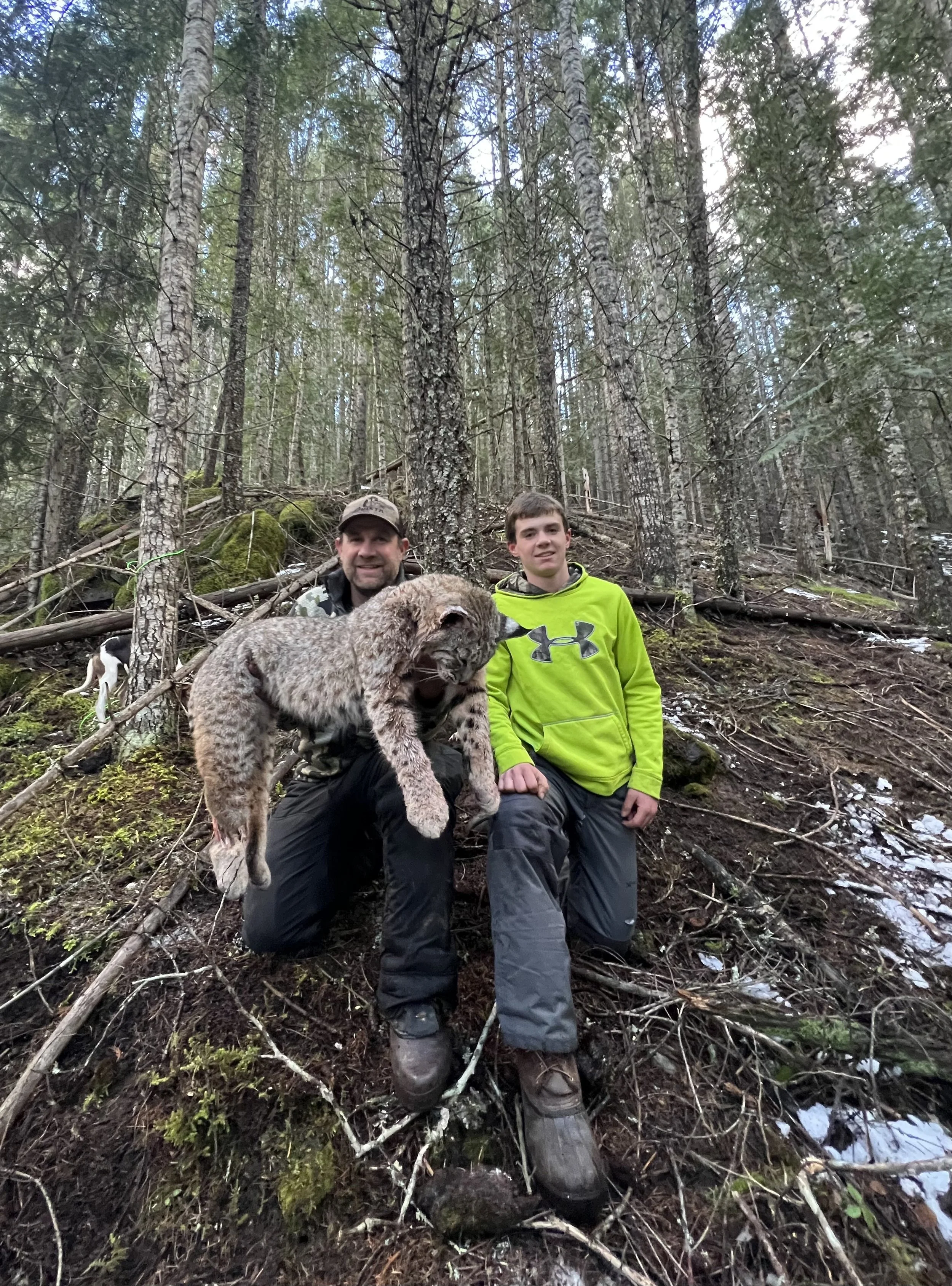 Two men in a forest with tall trees, one kneeling and holding a bobcat, the other standing next to him. The man holding the bobcat is smiling, and the other looks at the camera. The forest floor is uneven with moss, twigs, and snow patches.