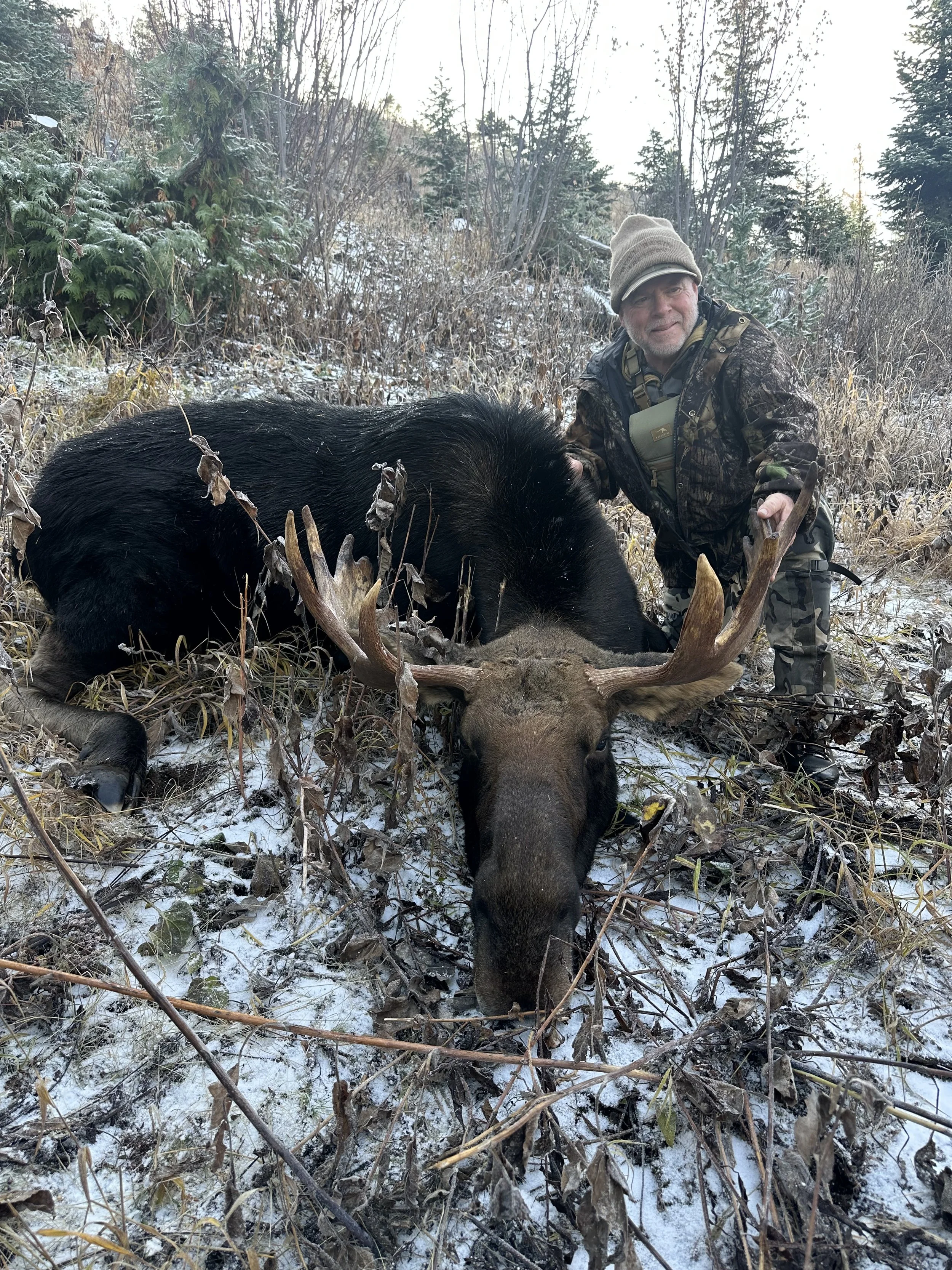 A man dressed in camouflage hunting gear kneeling next to a large dead black bear with prominent antlers, in a snowy, forested area.