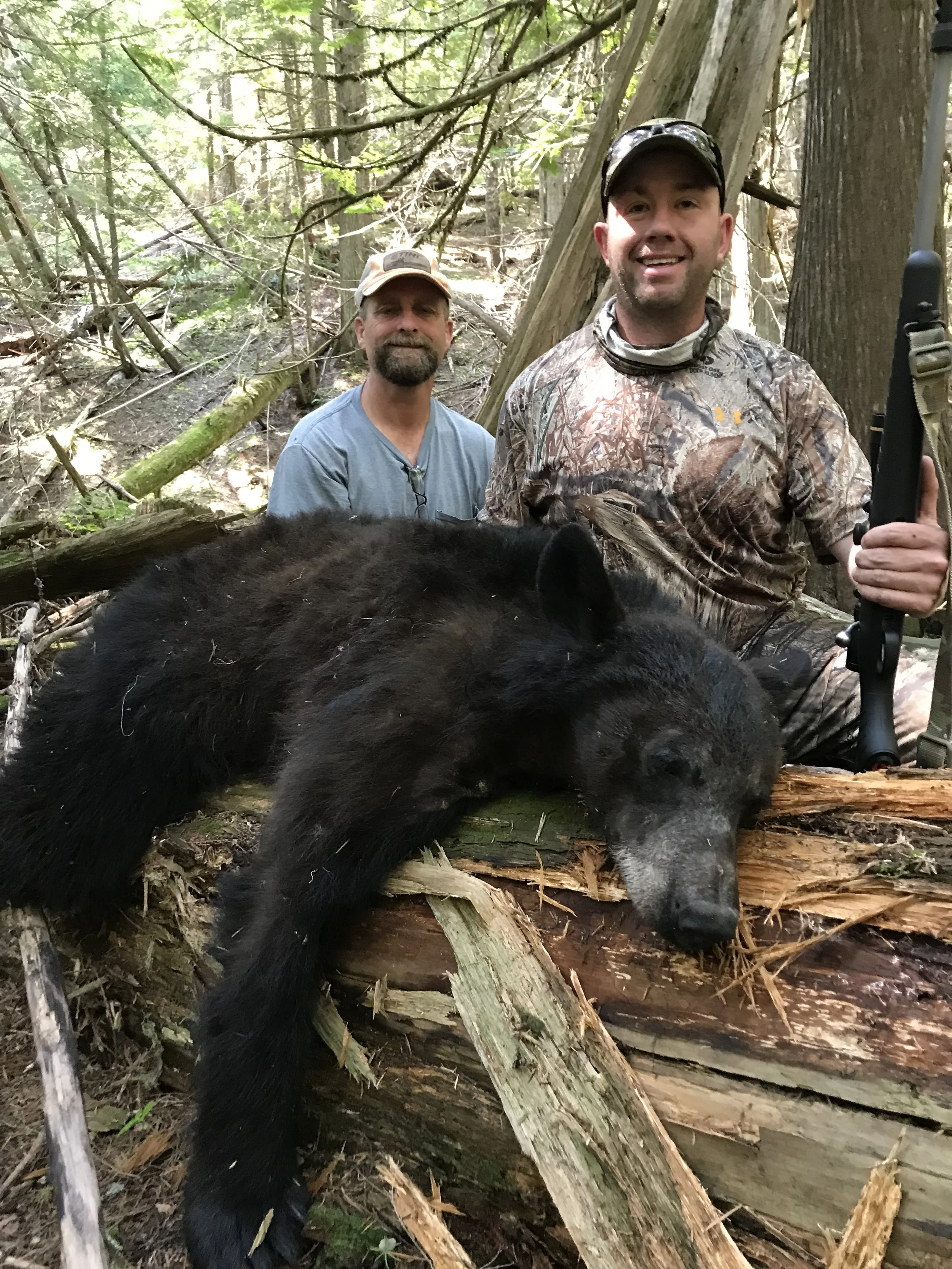 Two men in camouflage and outdoor clothing in a forest with a large, dead black bear lying on a fallen log in front of them.