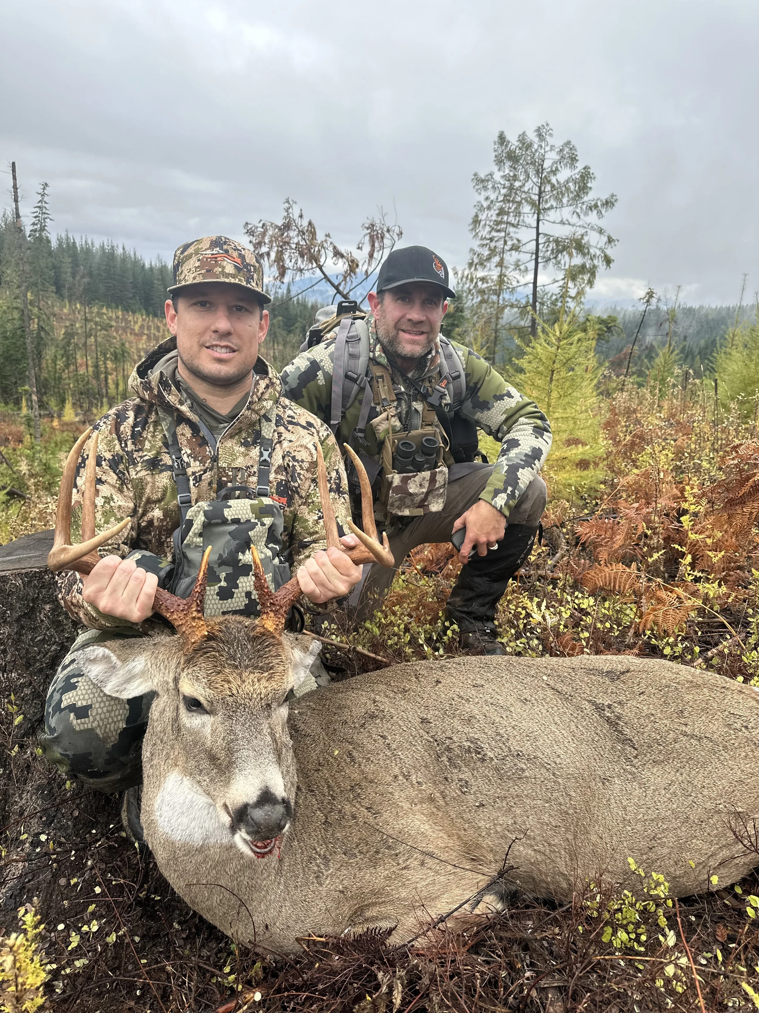 Two hunters in camouflage gear kneeling next to a large dead deer with antlers, in a forested area during overcast weather.