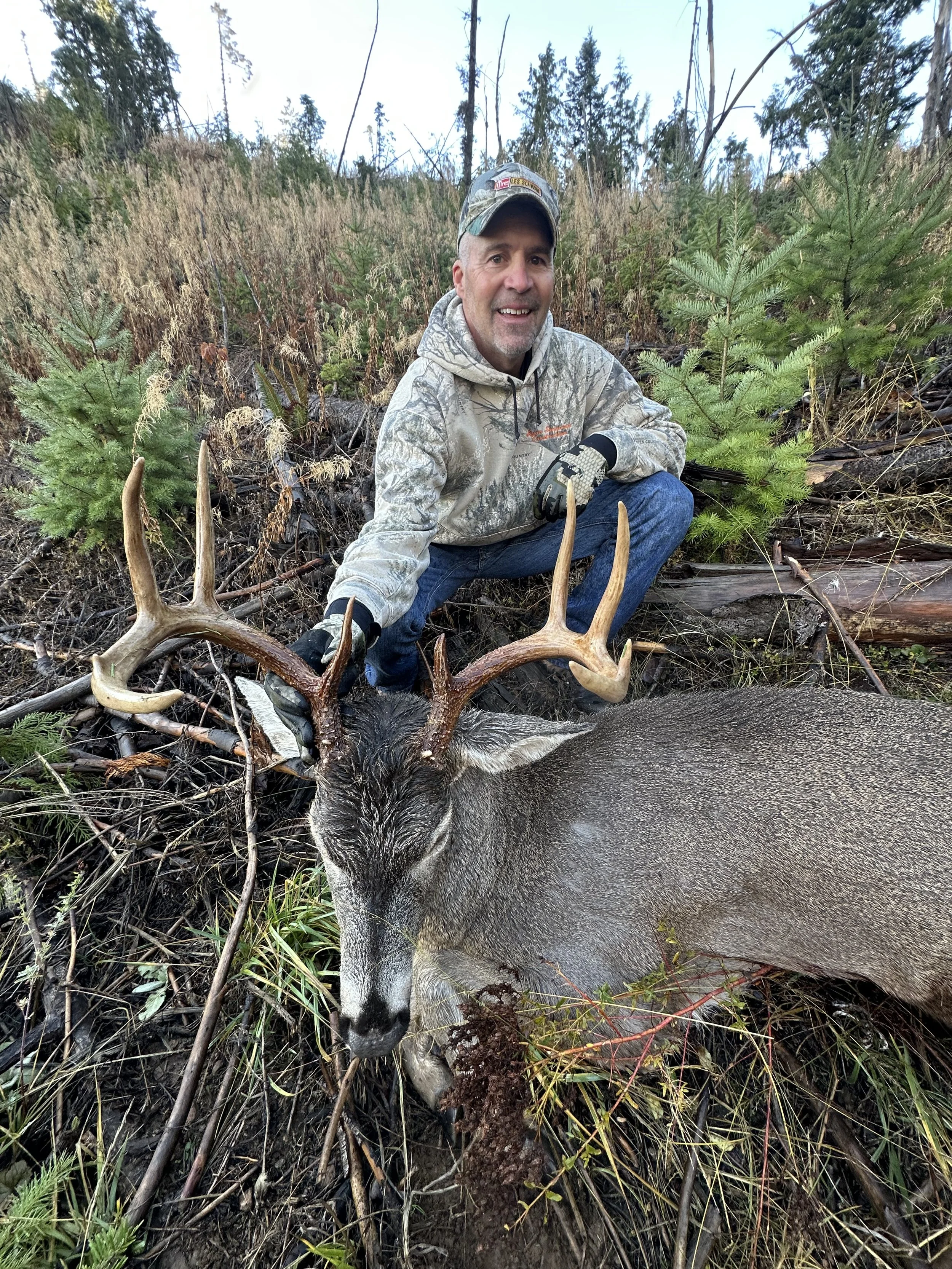 A man kneeling next to a large deer with prominent antlers in a forested area, smiling at the camera.