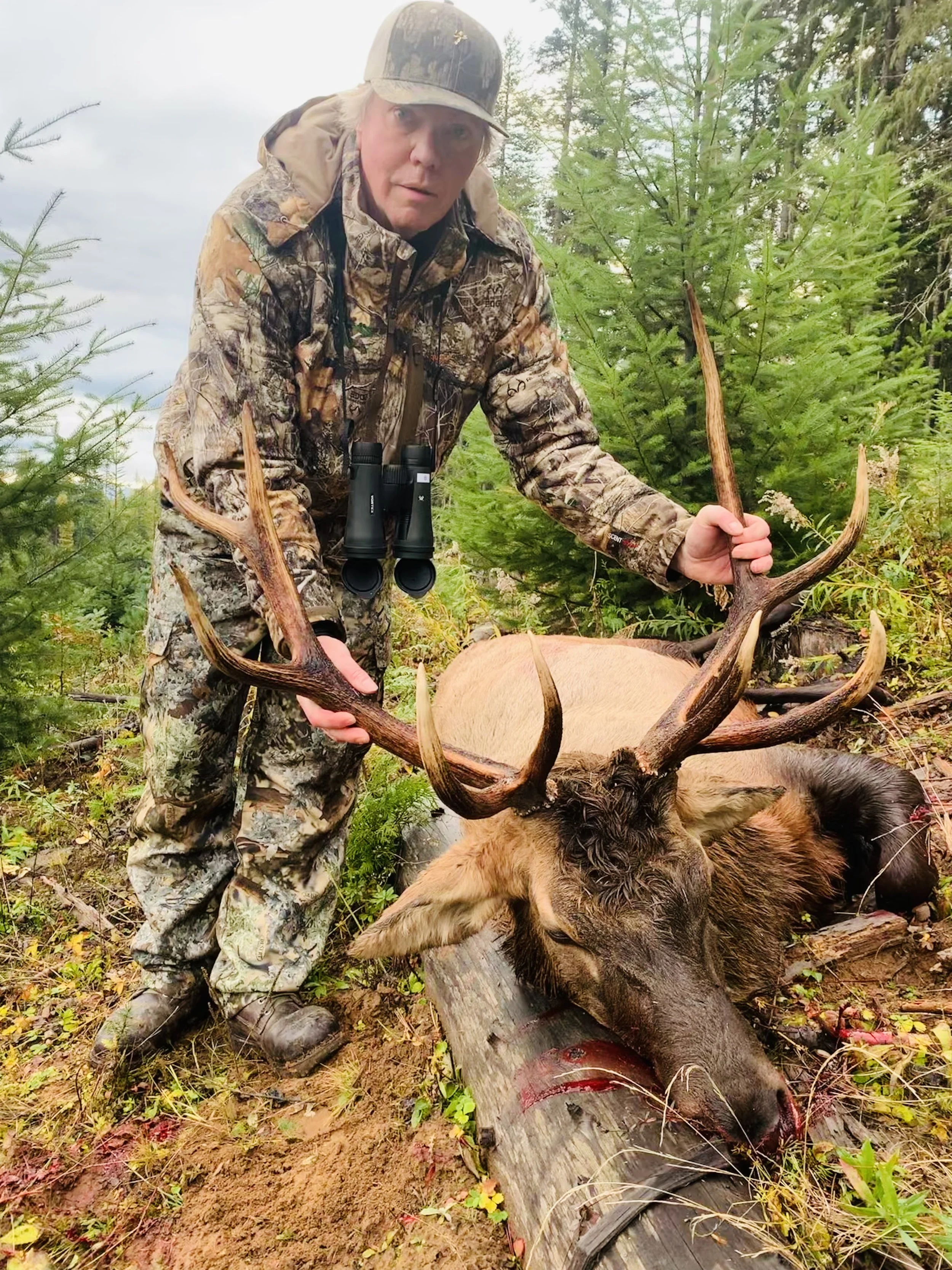A man wearing camouflage attire and binoculars around his neck is kneeling outdoors, holding large antlers of a recently hunted deer, with the deer lying on the ground. The scene is in a forested area with green trees and shrubs.