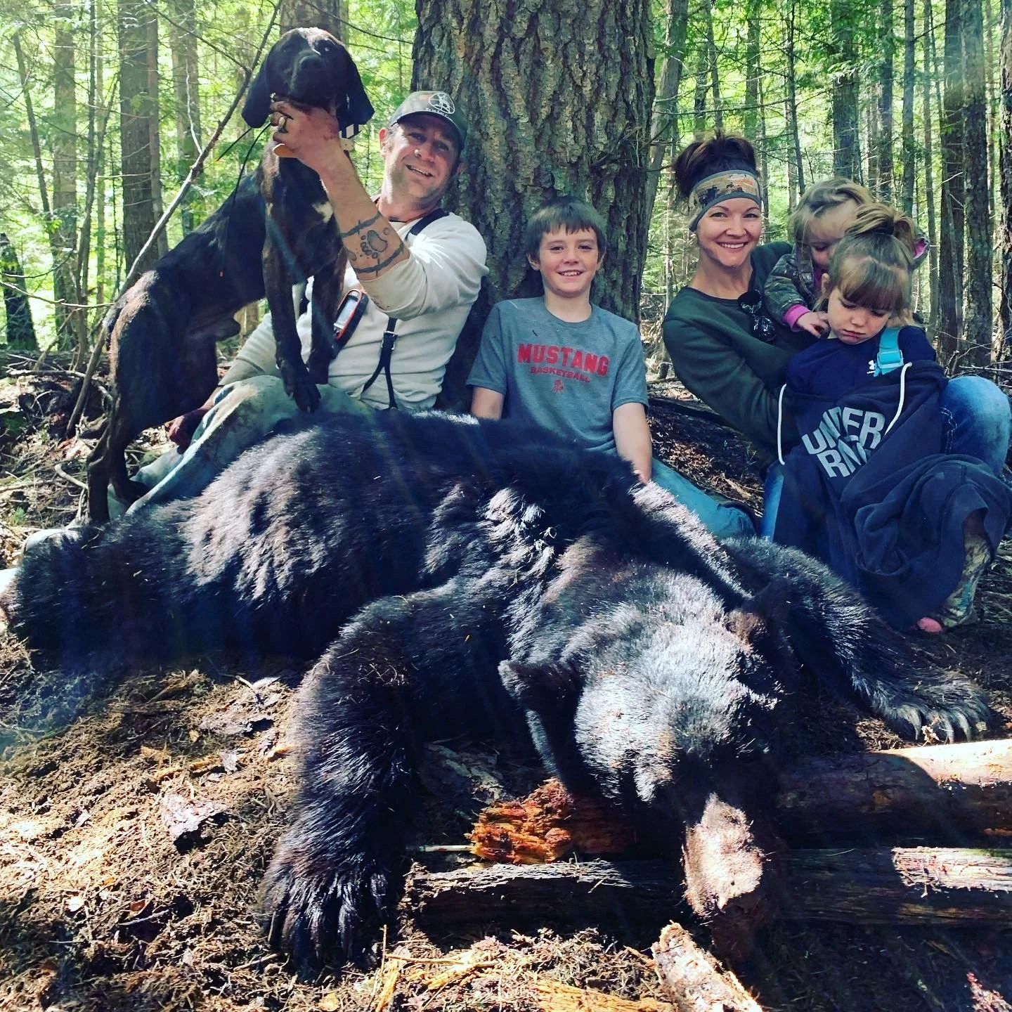 Group of five people, including children, in a forest with a large black bear and a hunting dog present. One adult man is holding a young black and white dog, and there is a large black bear lying on the ground.
