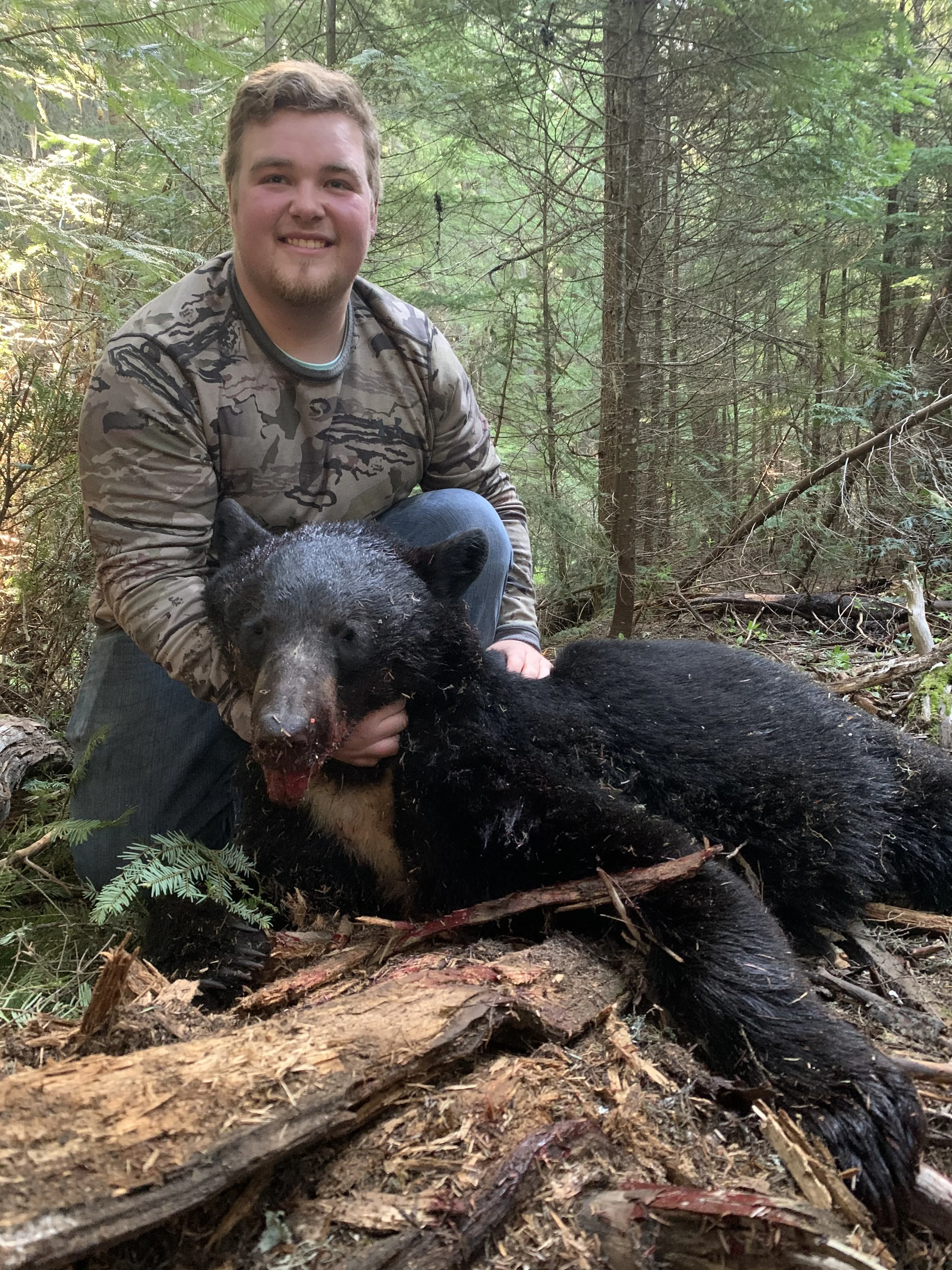 A young man kneels in a forest, smiling, while holding a freshly killed black bear on the ground.