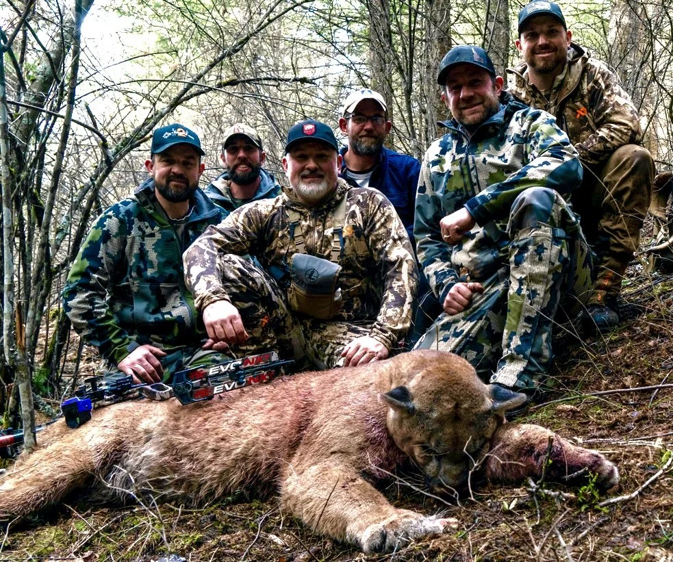 Six men in camouflage and outdoor gear pose behind a large, dead juvenile bear in a forested area. The bear is lying on the ground.