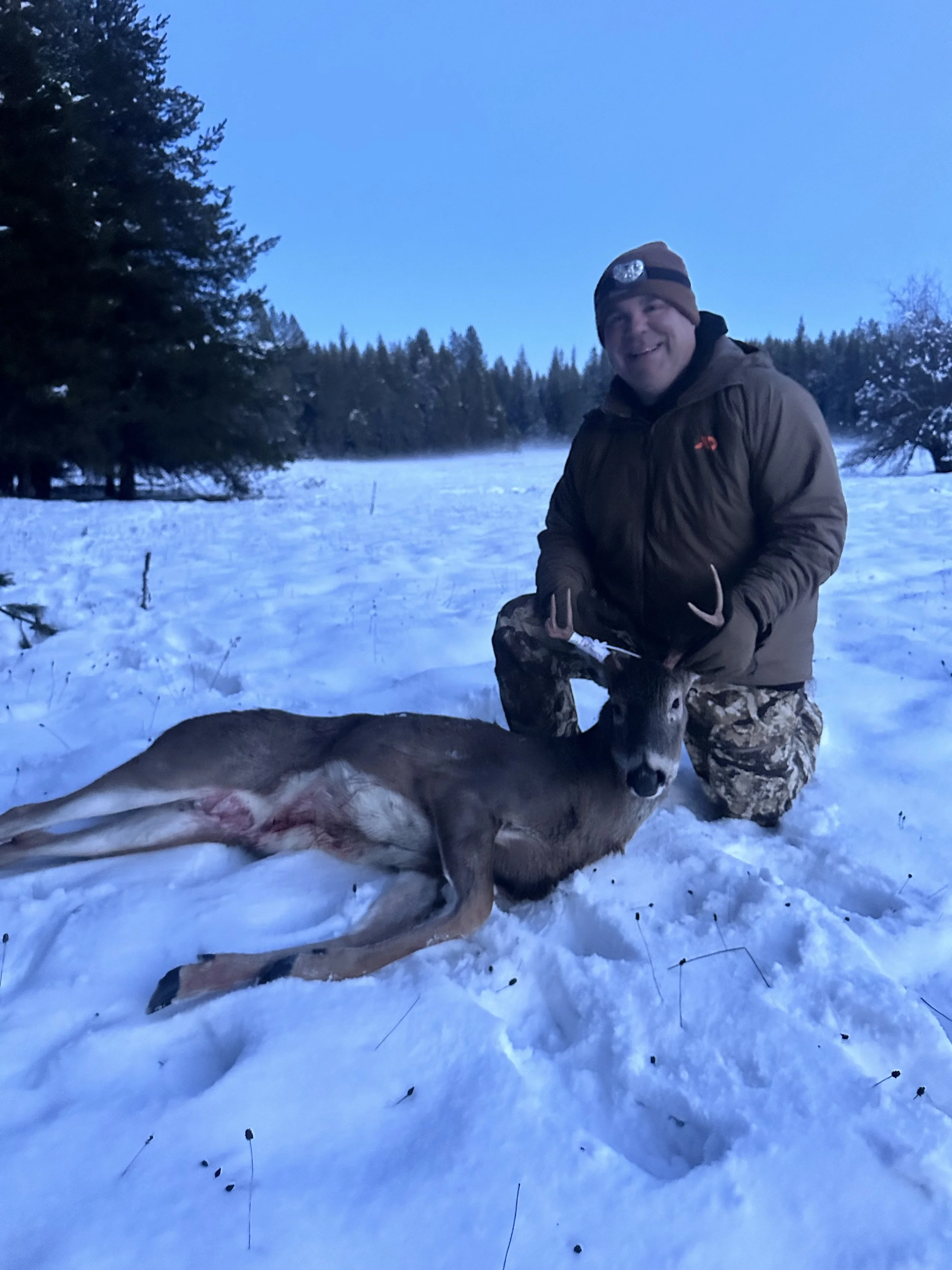 A man kneeling in snow holding a rifle next to a deer he has hunted, with trees and a cold landscape in the background.