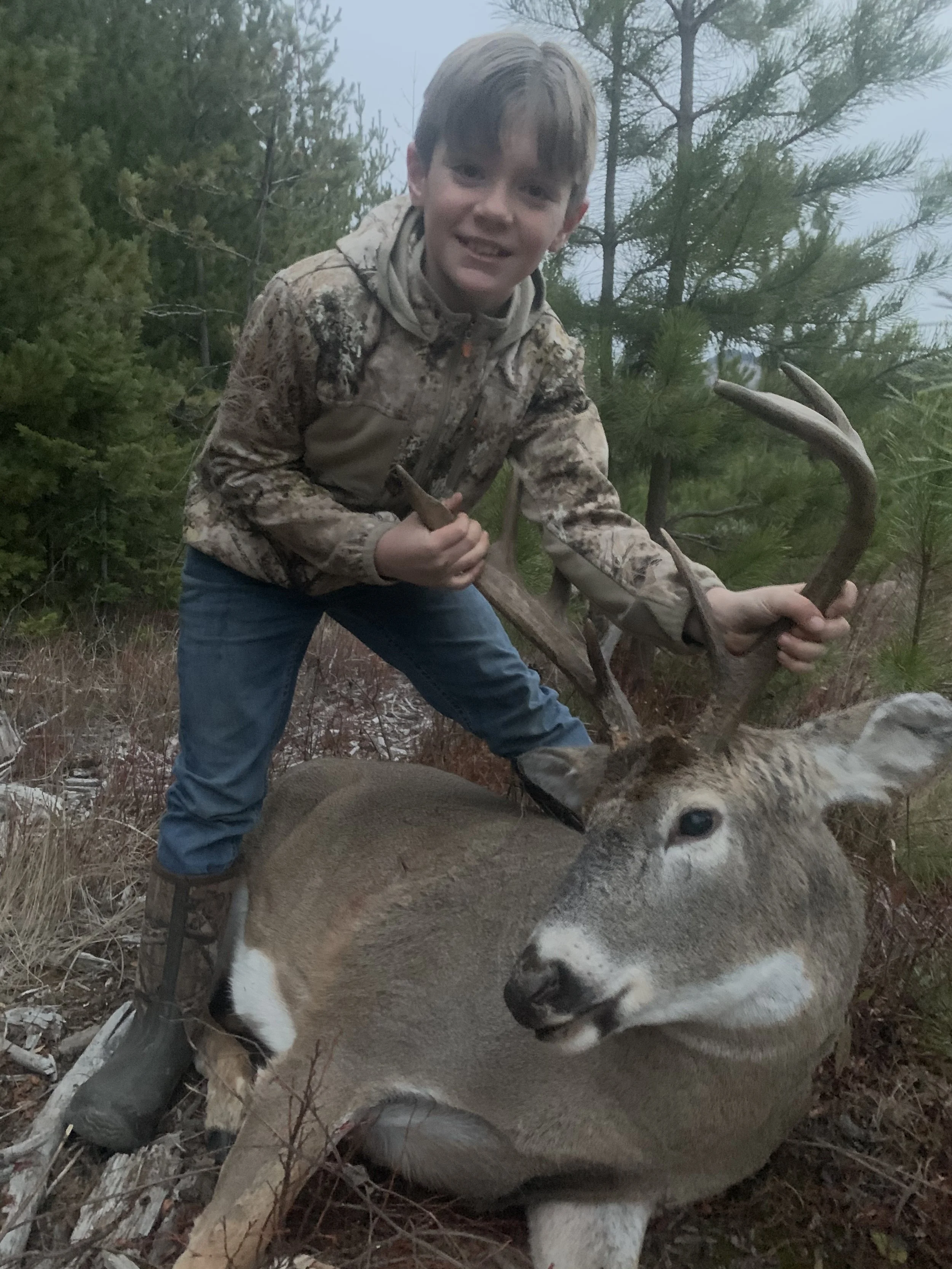 A young boy in camouflage jacket and blue jeans posing with a large deer he hunted, outdoors in a forested area.