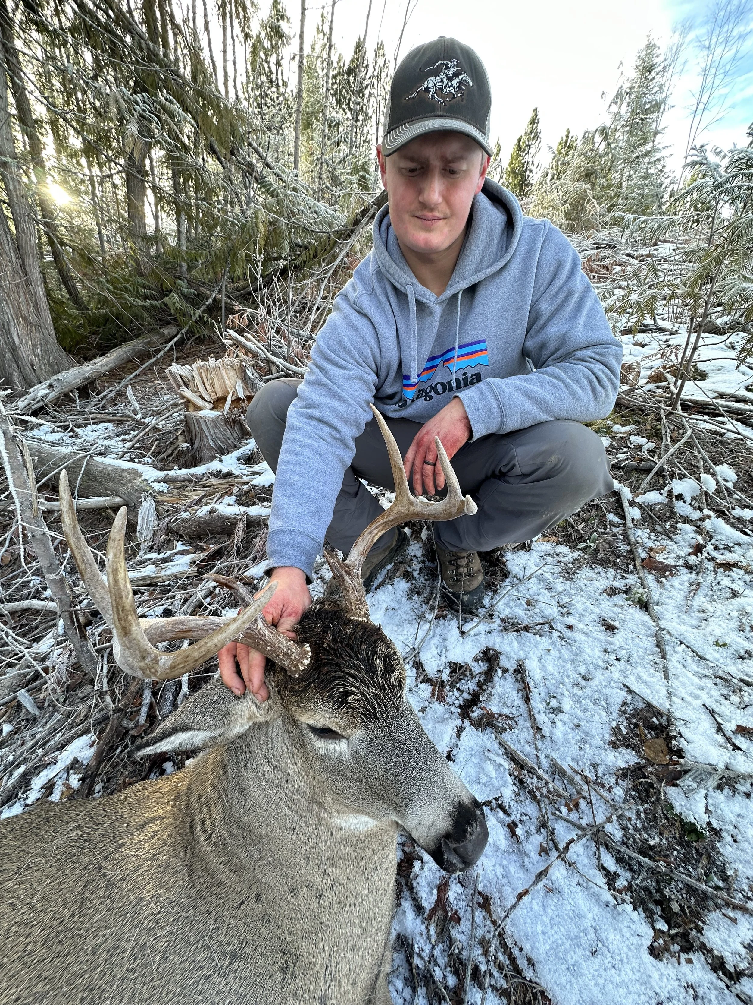 A young man in a gray hoodie and baseball cap squats in a snowy forest, holding the antlers of a harvested deer with a calm expression.