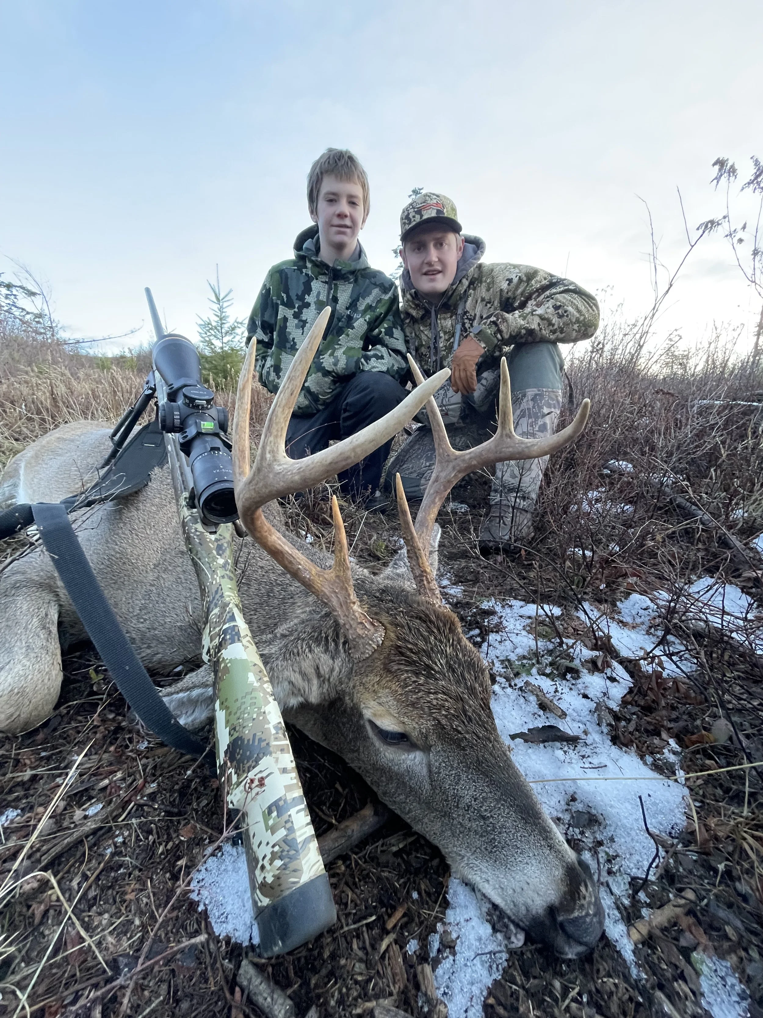 Two hunters pose behind a large deer with antlers, lying on the ground in a wooded area with snow; one with short hair, the other with a cap and camouflage jacket, with a camouflage rifle resting against the deer.