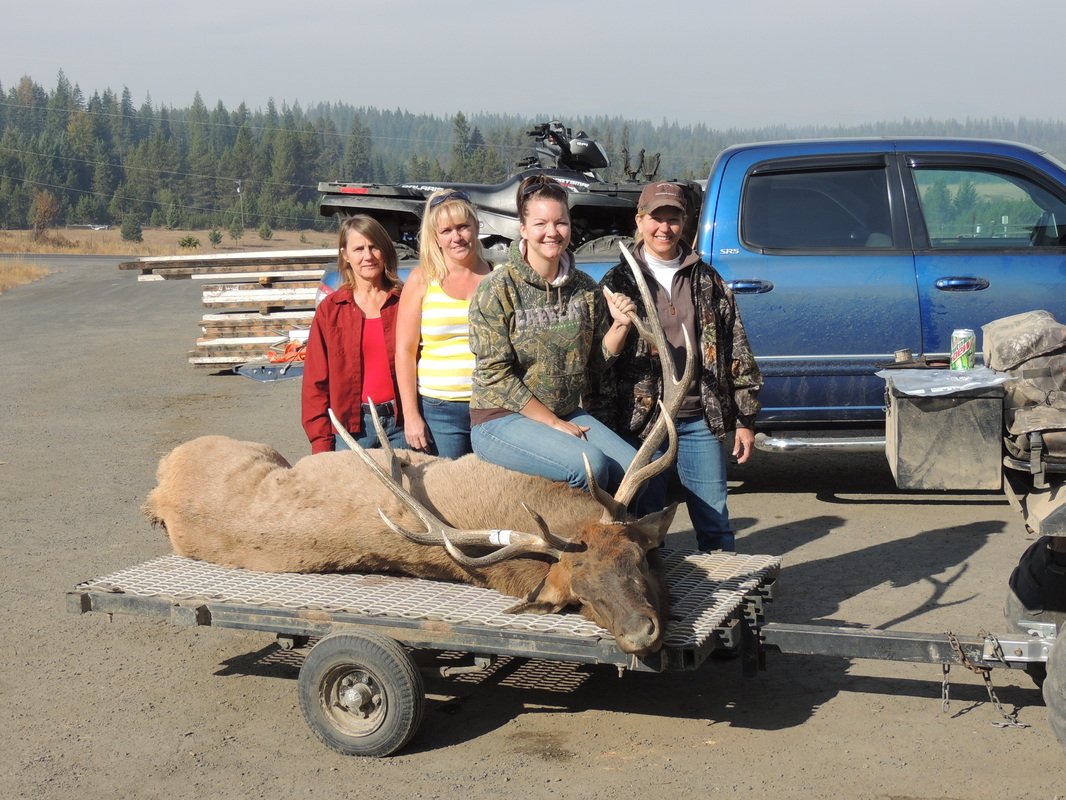 Four women standing behind a elk with large antlers on a trailer, in a rural outdoor setting, with a blue truck and off-road vehicle in the background.