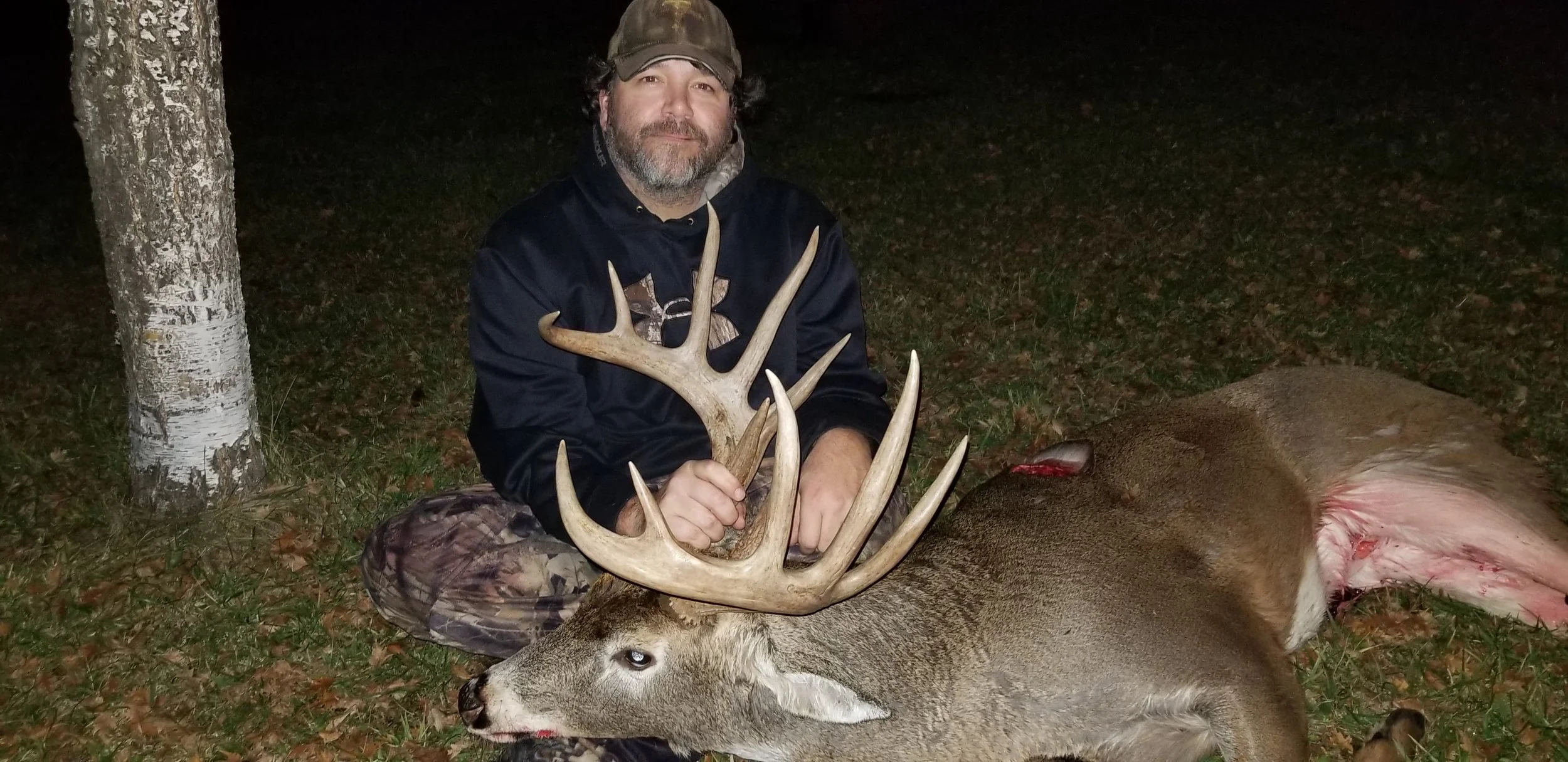 Man kneeling in camouflage pants and a black jacket, holding the antlers of a large deer with a white face and large antlers, lying on the ground next to a tree at night.
