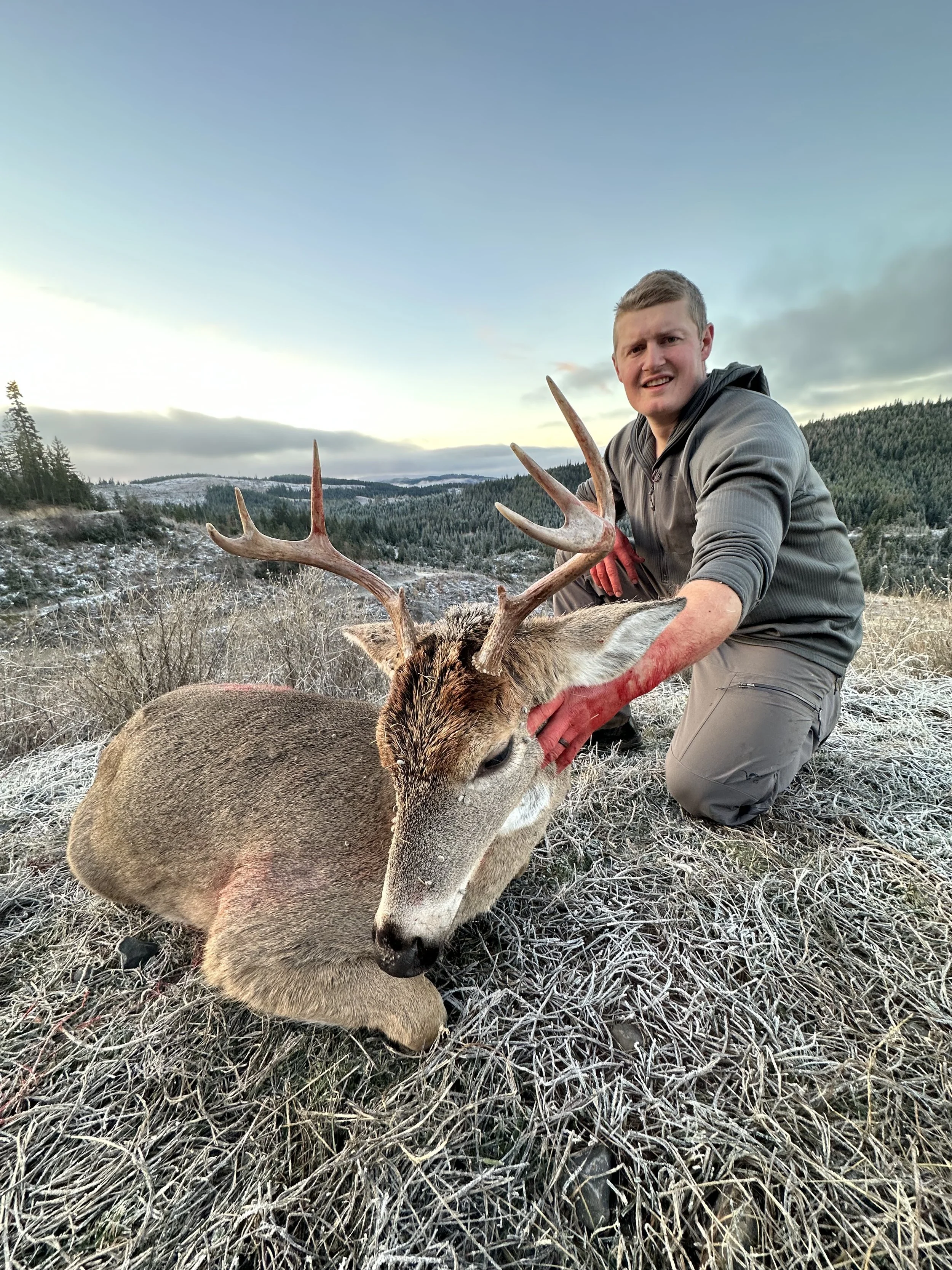 A man kneeling on frosty grass holding a large deer with antlers, outdoors in a mountainous landscape with trees and cloudy sky.