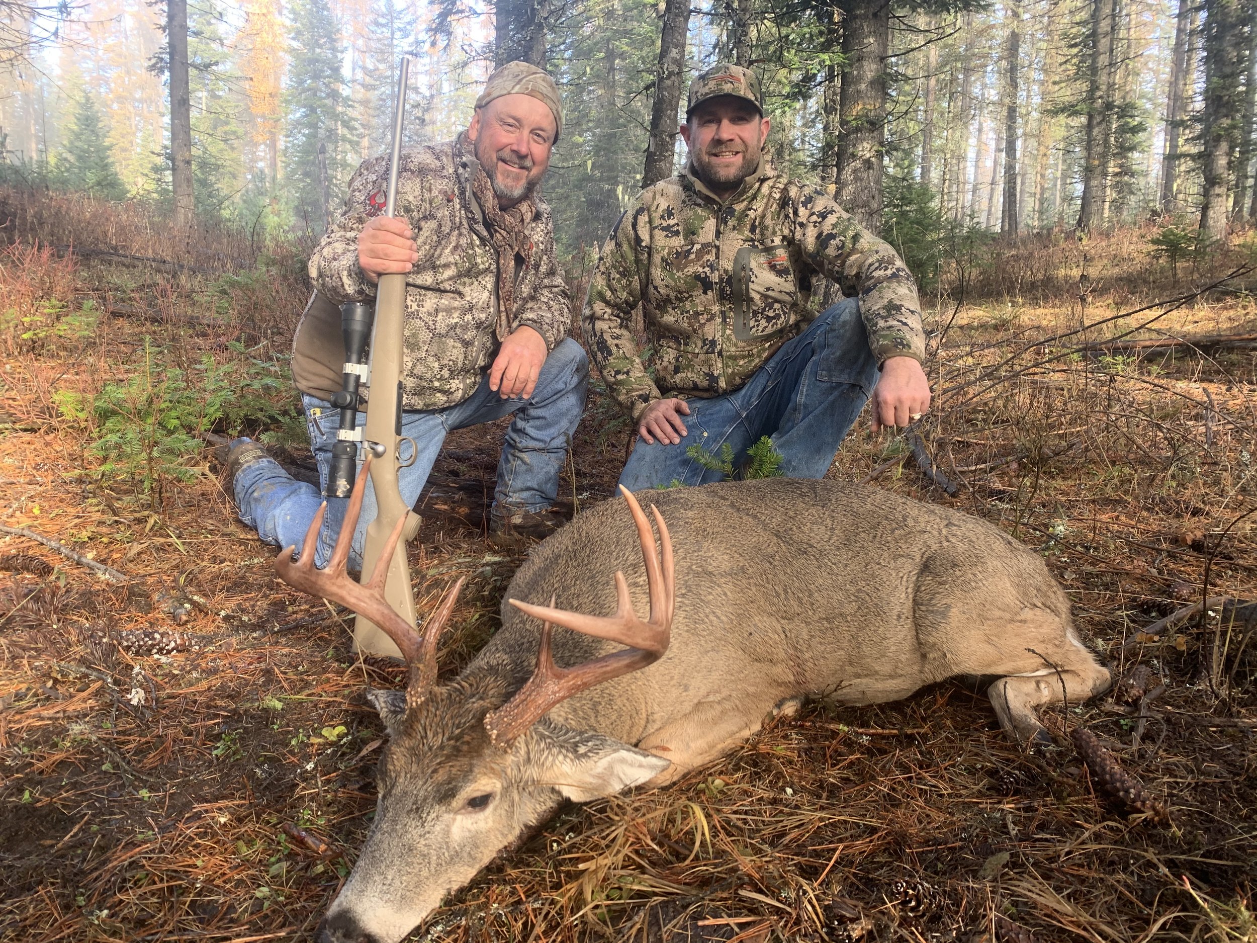 Two hunters kneeling next to a large dead deer with antlers in a forest. They are smiling and wearing camouflage clothing, with one holding a rifle.