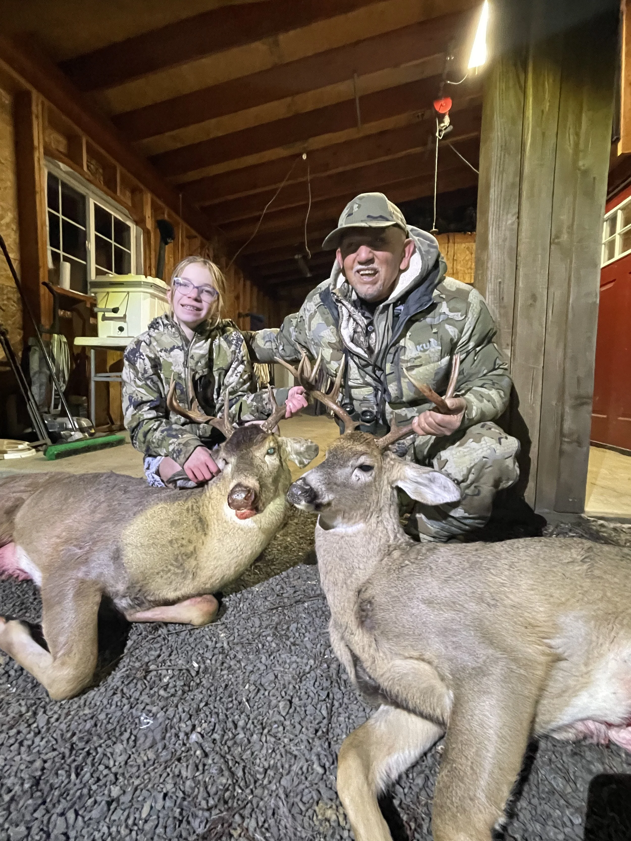 A young girl and an adult man in camouflage clothing kneel inside a wooden hunting shed, smiling and holding antlers from two recently hunted deer. The deer are lying on the ground, surrounded by gravel, with their eyes open. The shed has a window, t