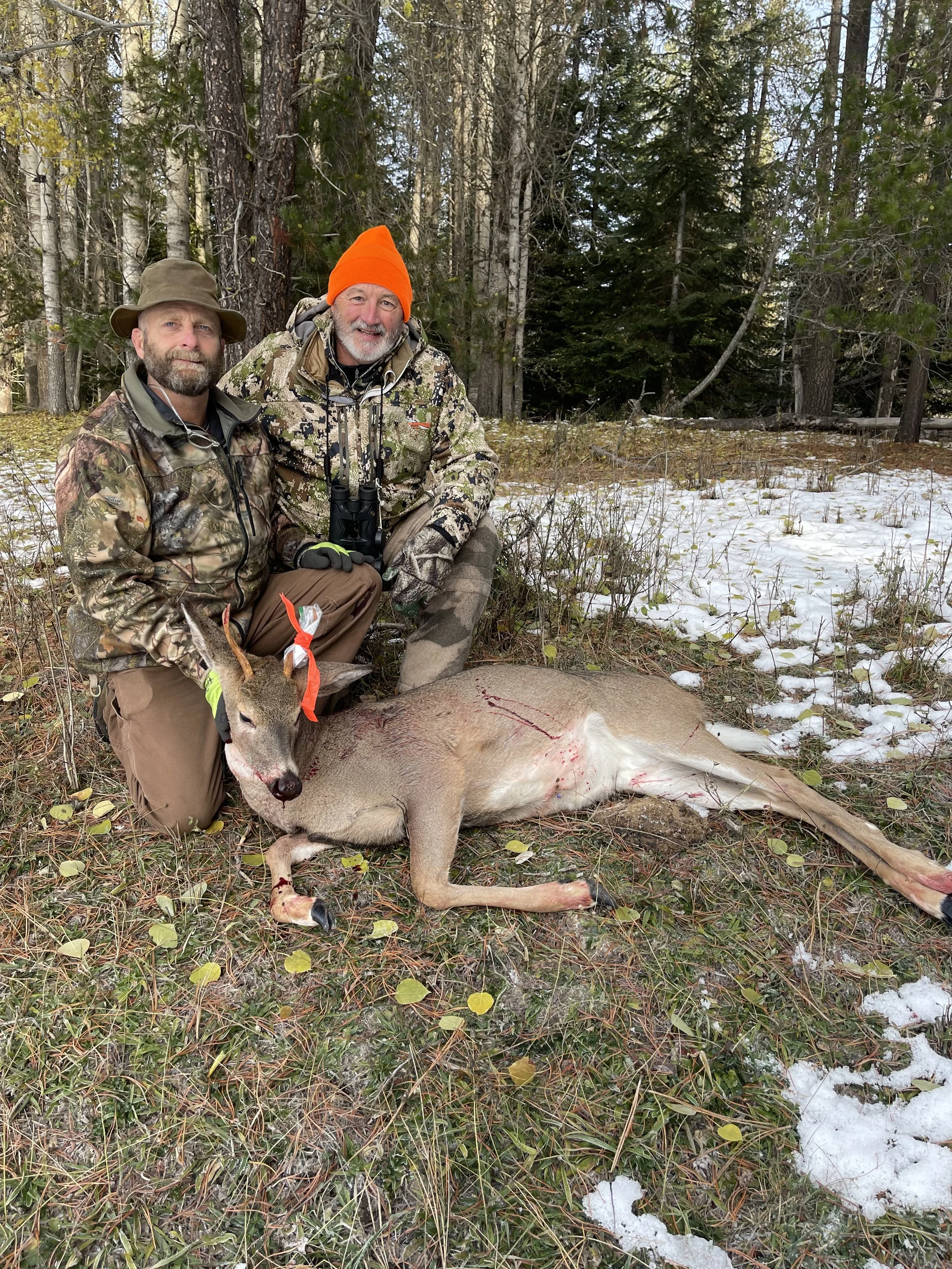 Two hunters kneeling in a forest with snow, posing next to a hunted deer, wearing camouflage clothing and outdoor gear.