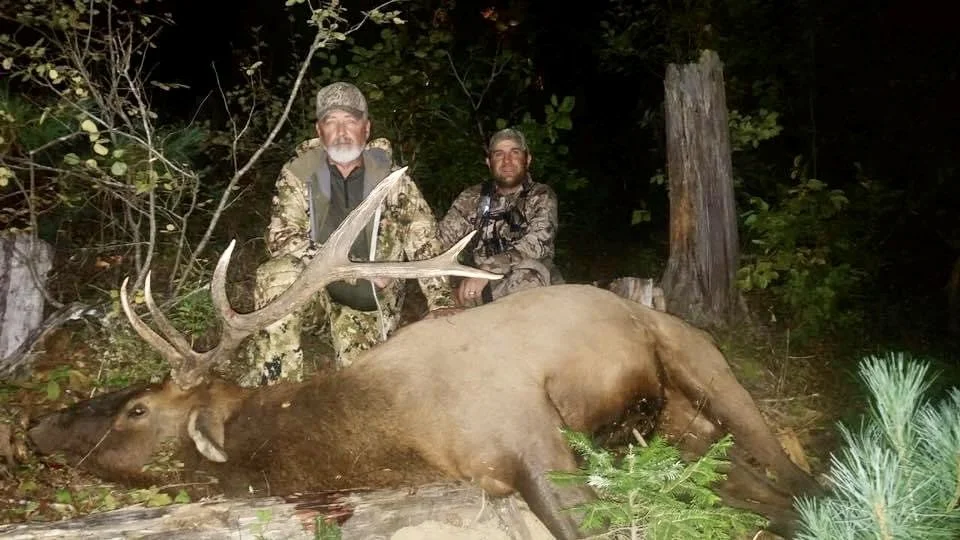Two hunters in camouflage gear kneeling behind a large elk carcass in a forest at night.