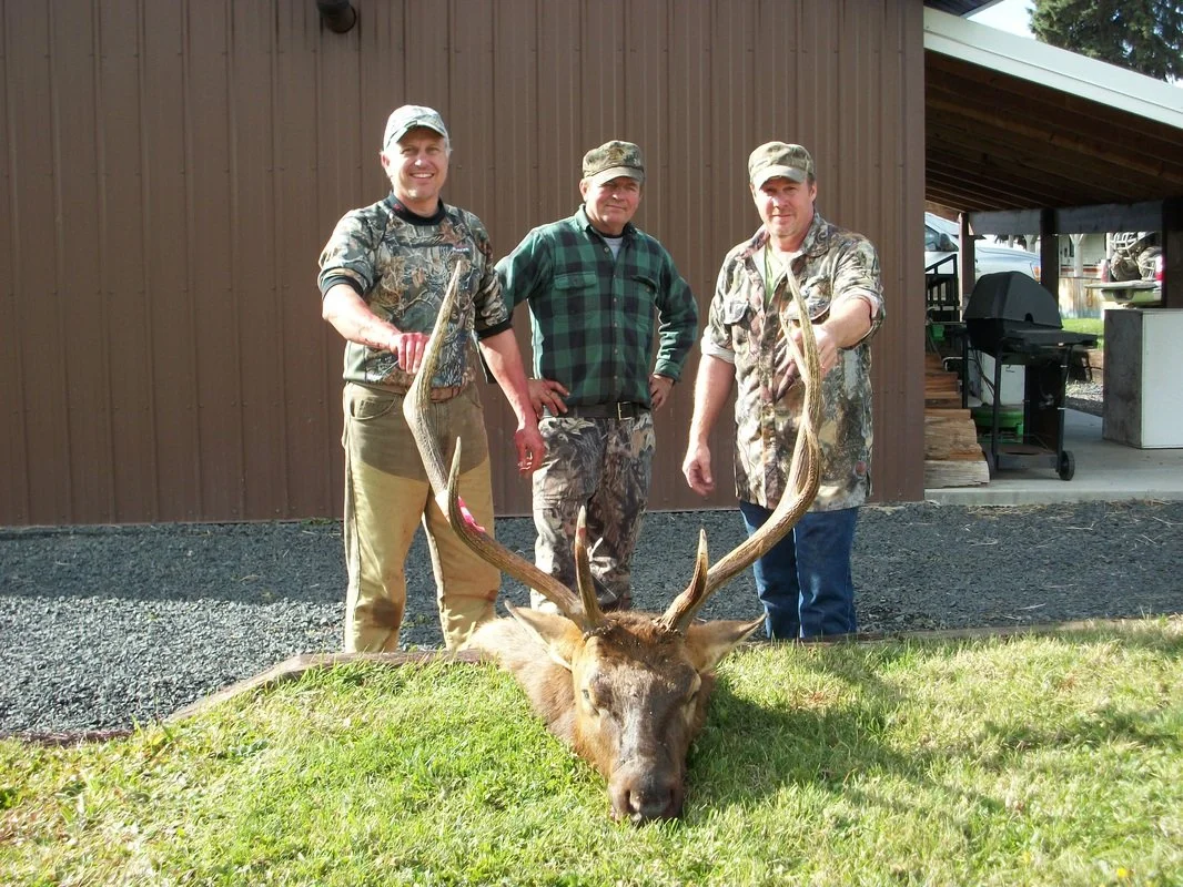 Three men stand outdoors behind a large deer with antlers lying on the grass. The men are wearing camouflage and plaid clothing, and the background shows a brown building and some outdoor equipment.