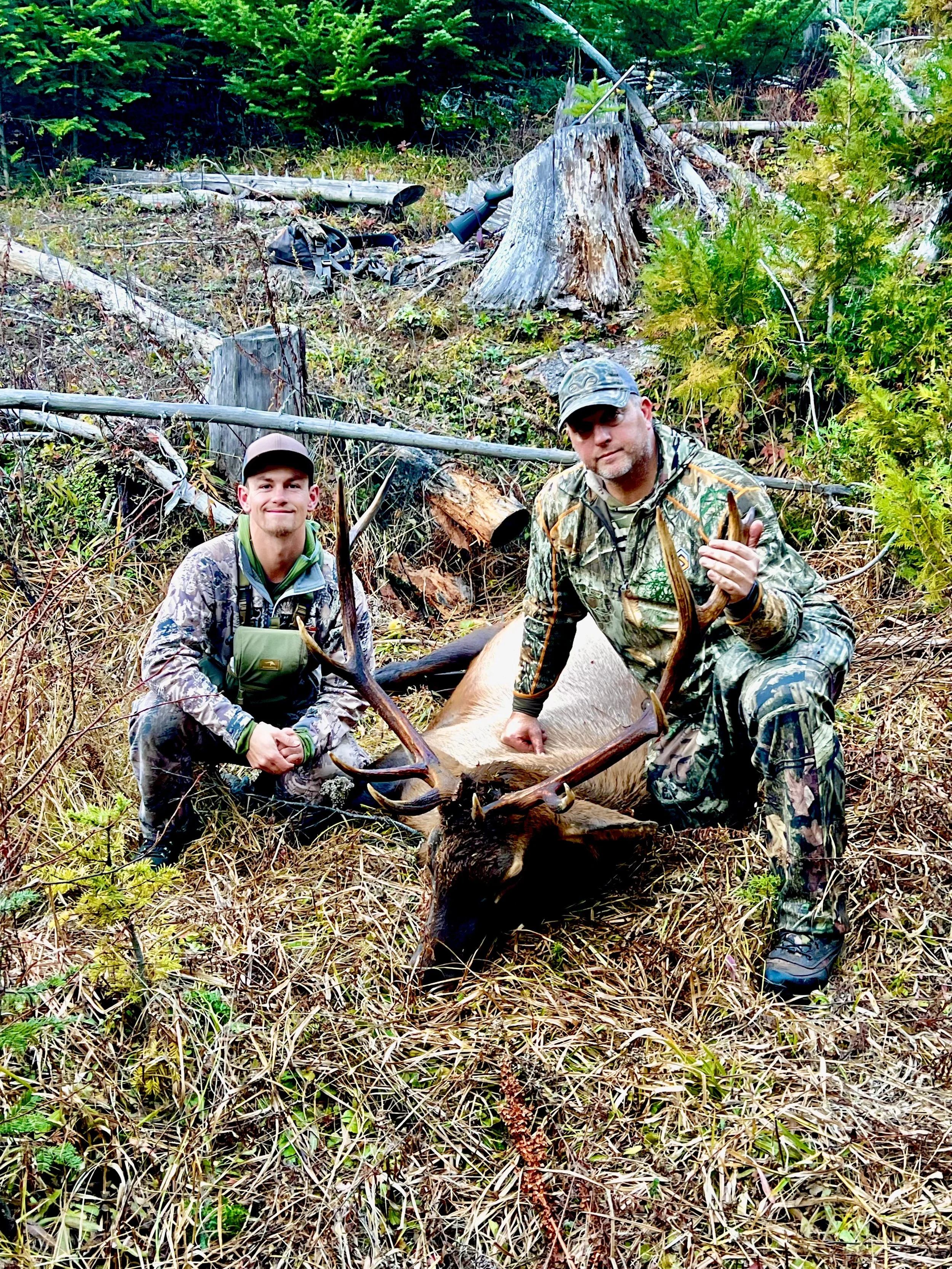 Two hunters in camouflage clothing kneeling beside a large dead deer with antlers in a forested area.